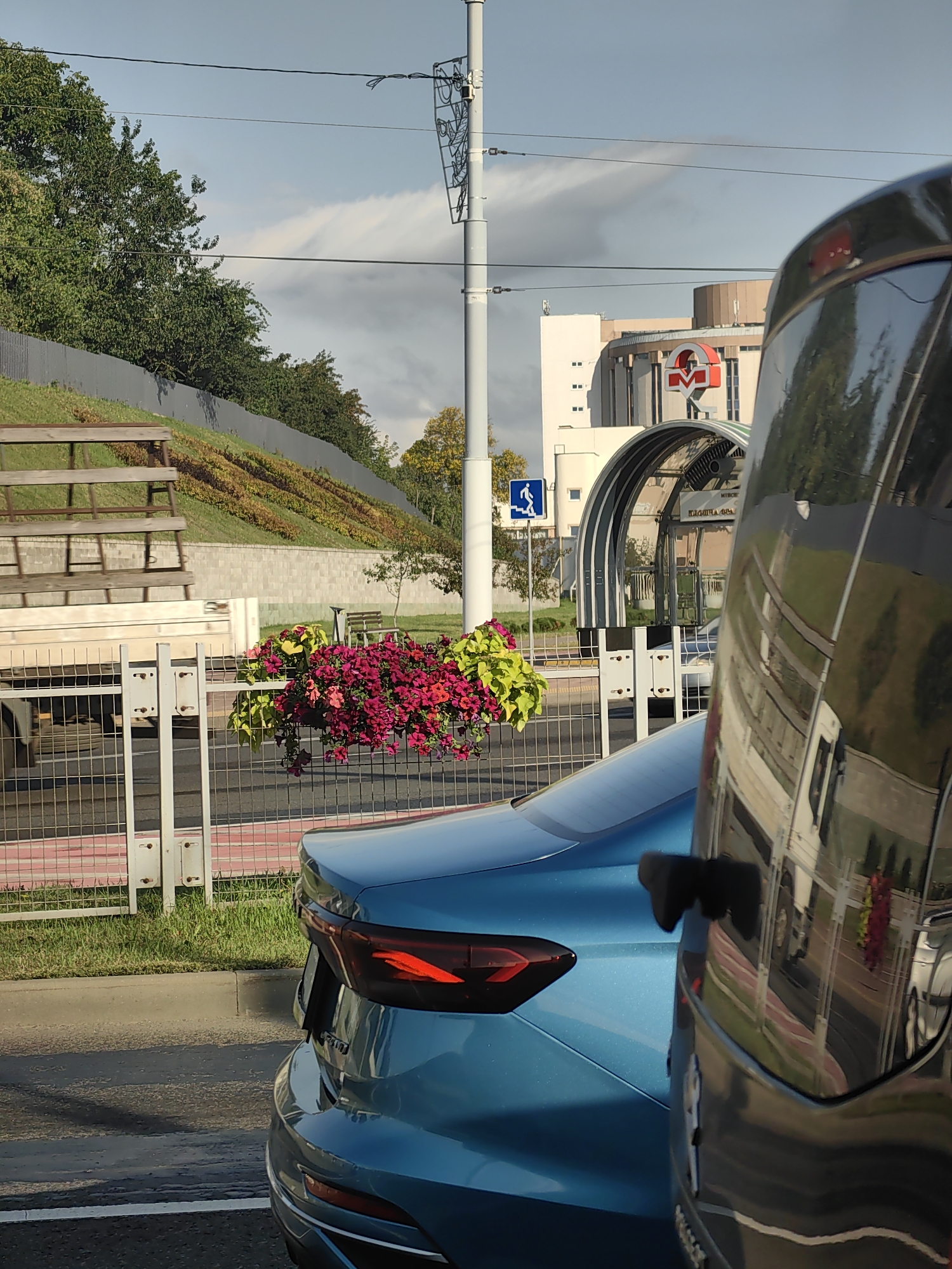 The image depicts a street scene in Minsk, Belarus, on a partly cloudy day. In the foreground, the rear of a blue sedan is visible, parked on an asphalt road. To the right, the reflective surface of another vehicle, possibly a van, partially obscures the view. Beyond the vehicles, a metal fence with a grid pattern separates the road from a sloped grassy area adorned with flowering plants in shades of pink and yellow. Further back, a modern building with a distinctive red and white metro logo is visible, along with an arched, transparent entrance structure to a metro station, identifiable by text that appears to read "ПЛОЩАДЬ ЯКУБА КОЛАСА" (Yakub Kolas Square). A blue pedestrian crossing sign with a white silhouette of a person walking is also present. The environment appears to be an urban setting with elements of public transportation infrastructure.