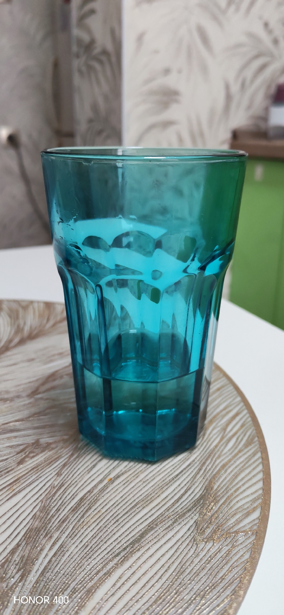 A blue glass with water is placed on a round decorative plate. The plate features a textured, feathered pattern in beige and white. The background is blurred, showing hints of a patterned wall and a green cabinet. The words "HONOR 400" are visible in the lower left corner of the image. The location and date are unknown.