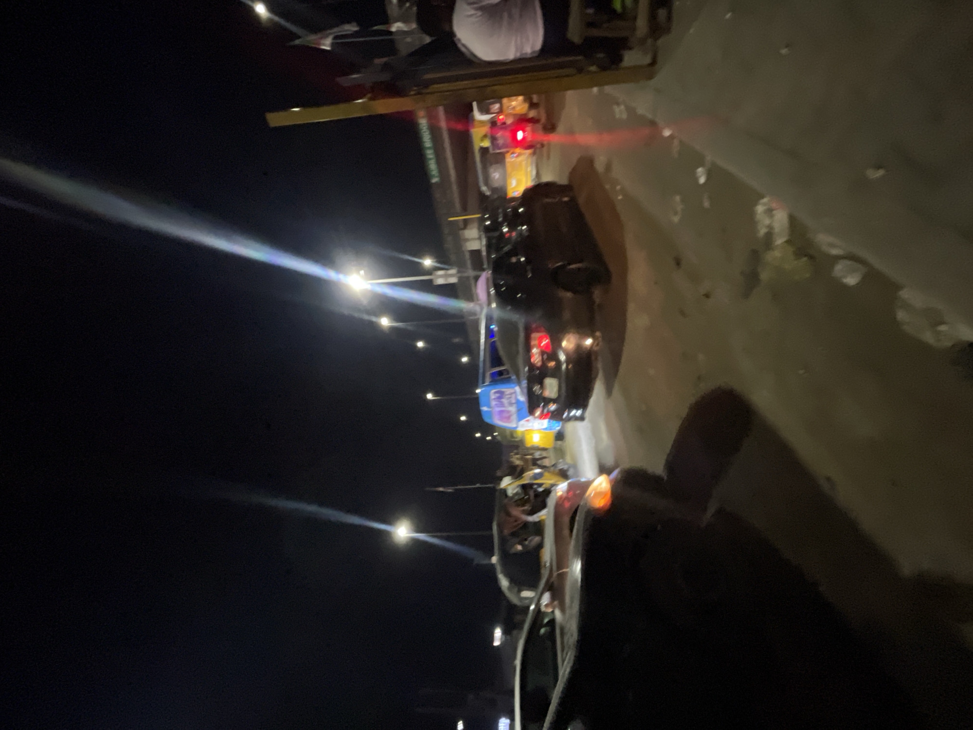 The image depicts a nighttime scene on a road in Lekki, Nigeria. Vehicles, including cars and possibly a commercial tricycle (Keke Napep), are visible on the roadway. Streetlights illuminate the area. A sign is visible, although the text is not entirely clear. The scene appears to be a standard traffic situation at night.