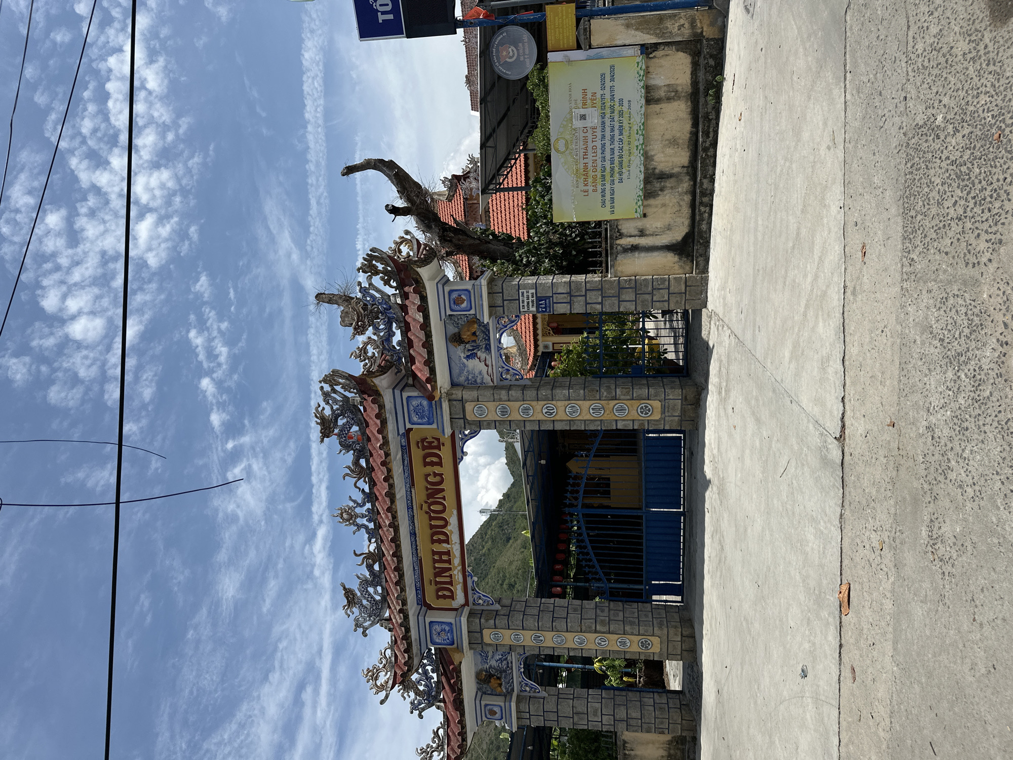 The image shows the entrance to the Dinh Duong De shrine in Bac Nha Trang Ward, Vietnam. The entrance is an ornate gate with a red tiled roof adorned with dragon sculptures. The gate is made of stone pillars and features blue metal gates. A yellow banner with Vietnamese text hangs on the wall to the right of the entrance. In the background, lush green mountains are visible under a partly cloudy blue sky. The ground in front of the entrance is paved with concrete. The scene is well-lit by daylight.