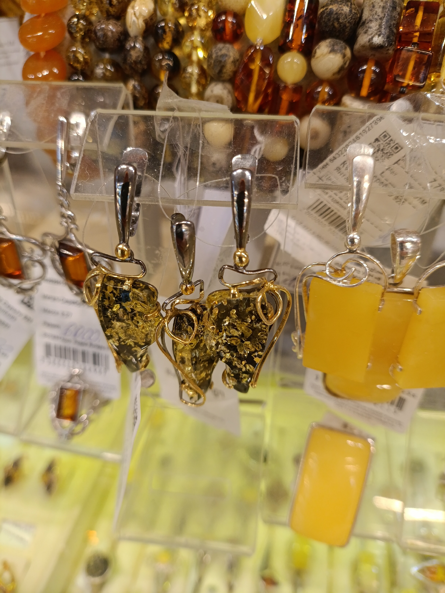 The image shows a close-up of jewelry, specifically earrings, displayed in a store. The earrings are hanging from a clear plastic display rack, with price tags visible on some of the items. The earrings feature amber stones of various colors, including dark amber with gold inclusions and bright yellow amber. The metal components of the earrings appear to be silver or a silver-colored alloy. Background elements show other jewelry items, including necklaces and pendants, also featuring amber. The lighting suggests an indoor environment, likely a jewelry store or market stall. This type of amber jewelry is typical for the city of Svetlogorsk, Russia, which is known for amber products.