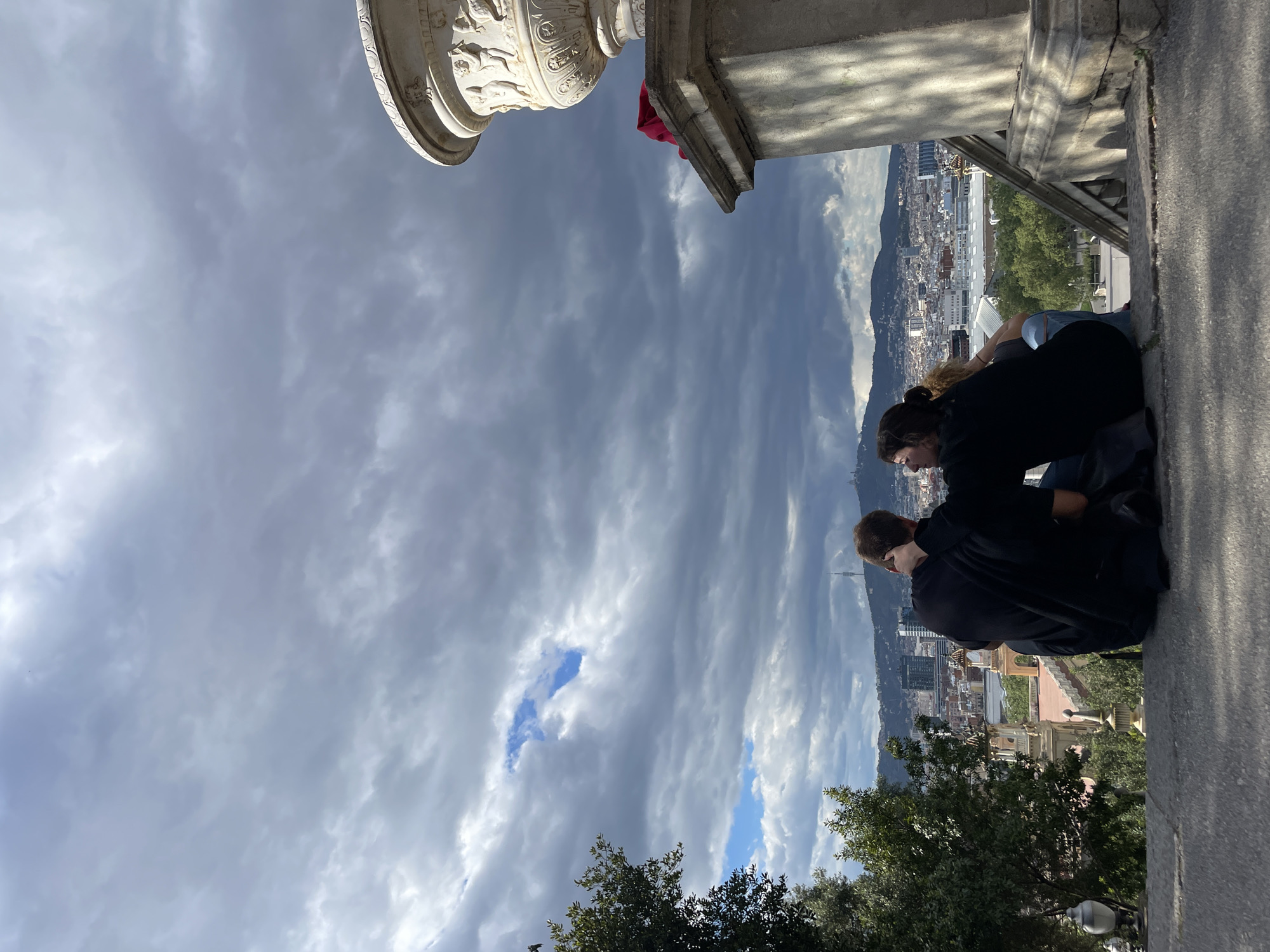 Two people are leaning over the ledge of a stone structure, looking out over a city. The city is likely Barcelona, Spain, given the distinctive urban layout and the mountainous backdrop. The foreground includes trees and a segment of the stone structure, adorned with classical carvings, suggesting a high vantage point such as a monument or overlook. The sky is predominantly cloudy with patches of blue.