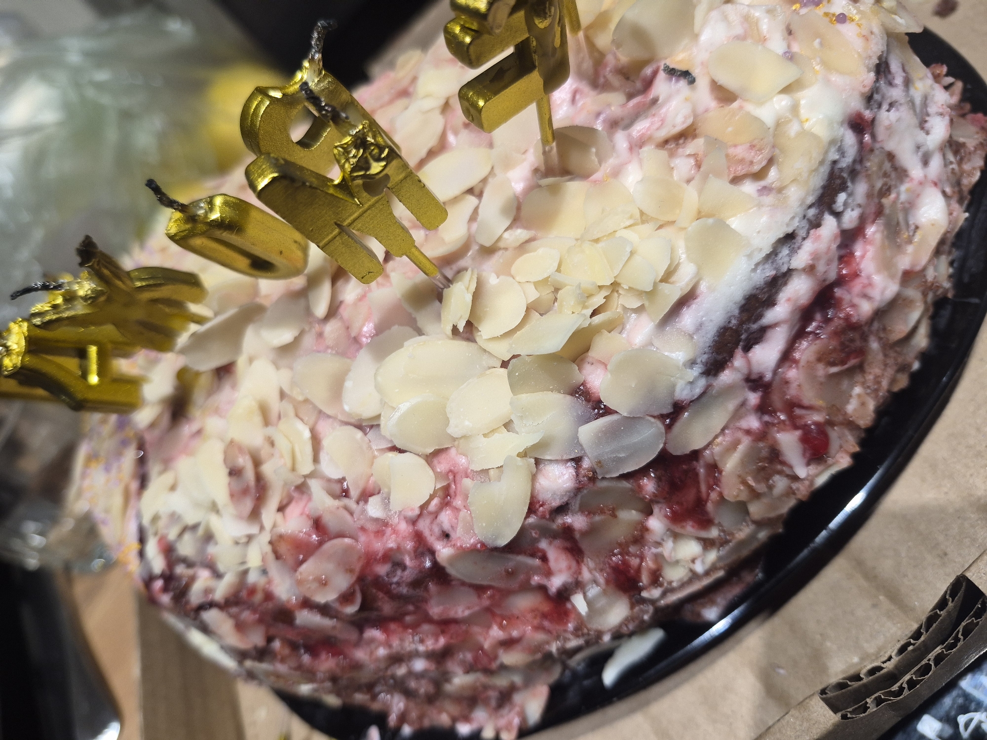 A close-up shot depicts a cake with almond slivers and red jam, decorated with gold "Happy Birthday" candles, likely indoors, with a blurred background. No specific details suggest the location is Chaikovsky, Russia. No people are visible. A piece of cardboard is visible in the foreground.