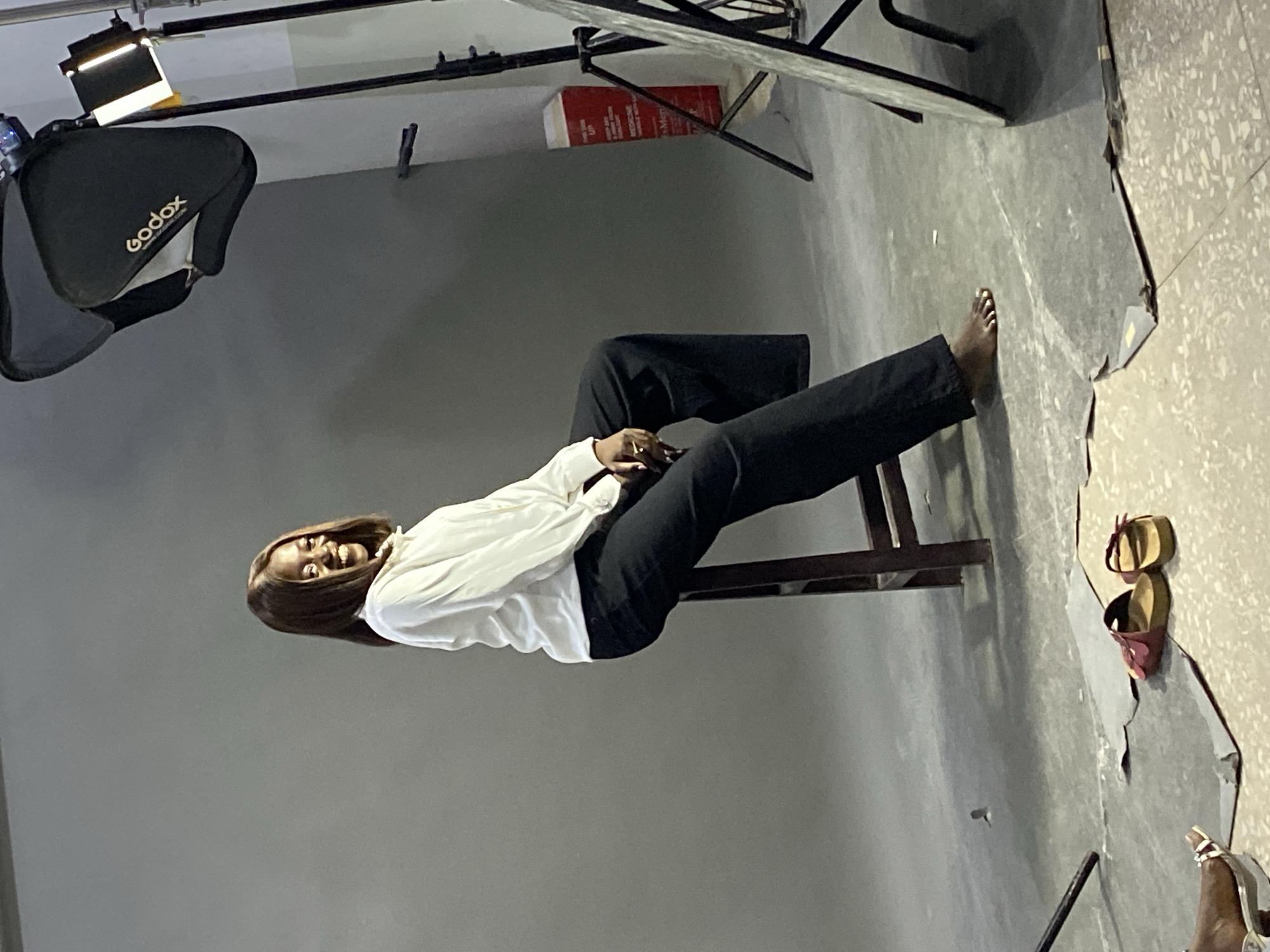 A woman with brown hair, wearing a white blouse and black pants, is sitting on a chair in a photography studio. The studio has a gray backdrop and the floor is gray with a light splatter pattern. Studio lighting equipment, including a softbox with the "Godox" brand name, is visible. A copy of "Step-Up Medicine" is on a light stand behind the seated woman. A pair of shoes is on the floor to the right of the woman. The woman is smiling. No information to indicate the specific location within the city of Jos, Nigeria is visible in the image.