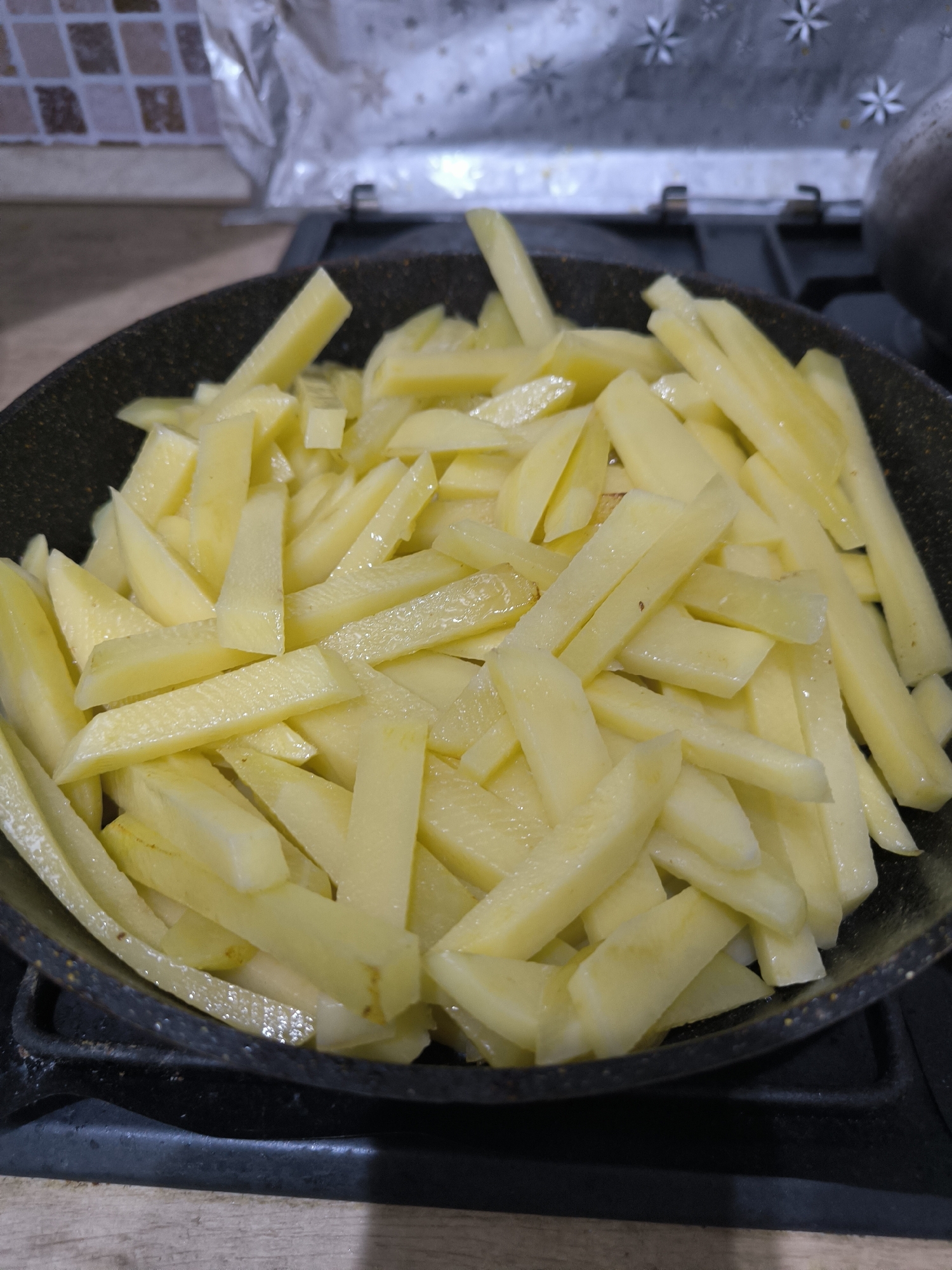 The image shows a close-up, overhead view of a frying pan filled with thinly sliced, raw potatoes. The potatoes are light yellow and appear to be coated in a light sheen of oil or water. They are arranged in a pile that fills most of the pan, with some overlapping. The pan itself is dark and appears to be non-stick, with a speckled texture on the interior. The pan is sitting on a black stovetop. In the background, out of focus, is a kitchen backsplash with a tiled pattern and a metallic material with a starry design. The lighting suggests an indoor setting, likely a kitchen. There is no discernible location in Chișinău, Moldova visible in the image.