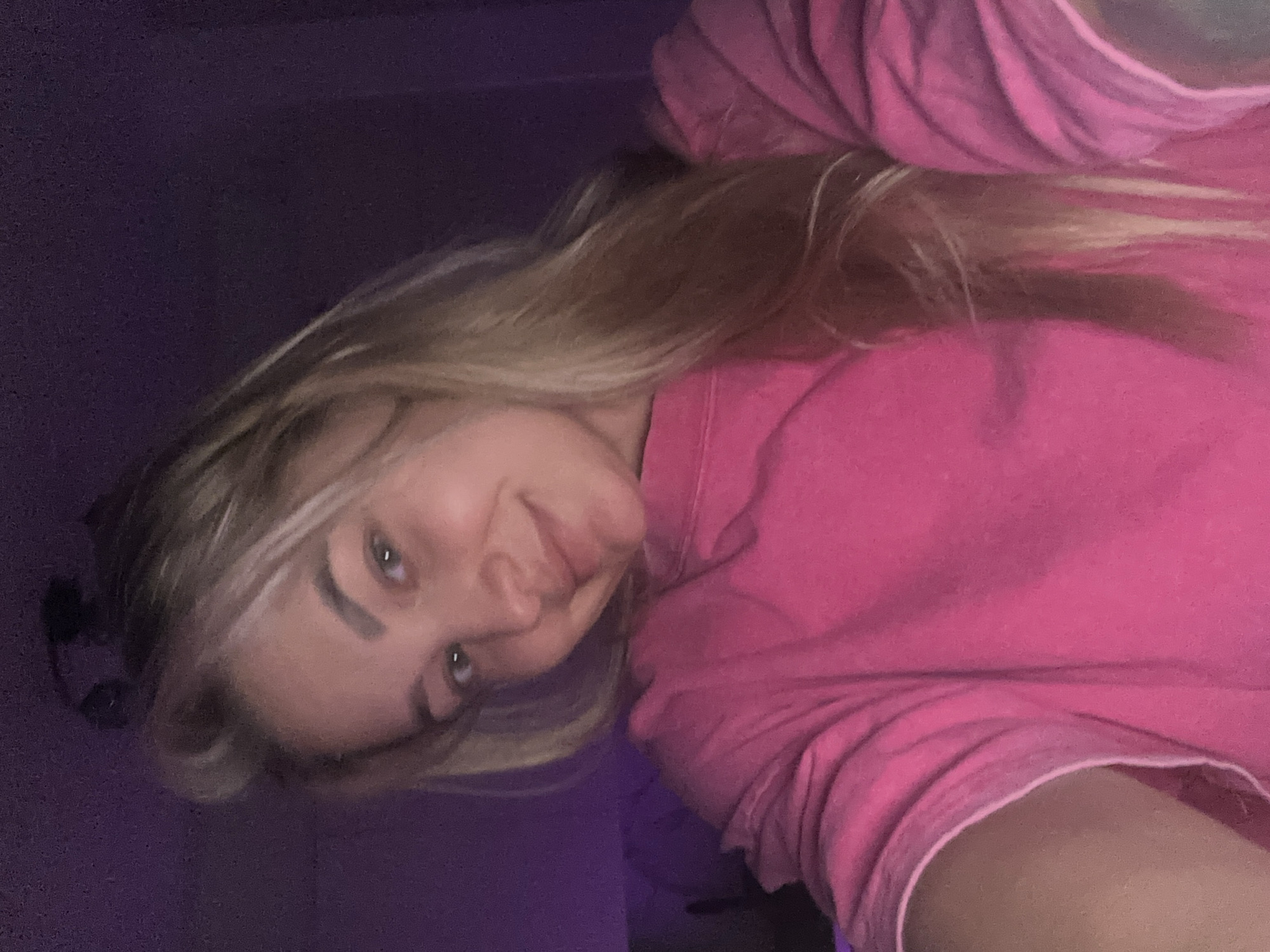 A close-up selfie features a young woman with long, blonde hair and light-colored eyes. She is wearing a vibrant, fuchsia pink hooded sweatshirt. Her head is tilted slightly, and she looks directly at the camera with a subtle, gentle smile or smirk, conveying a soft or playful emotion. Her eyebrows are well-defined. The background is a solid, dark purple or indigo hue, suggesting an indoor setting. The lighting is dim, and the image has a slightly grainy quality. The provided location context for this media is Budva, Montenegro. No visible text is present in the image.