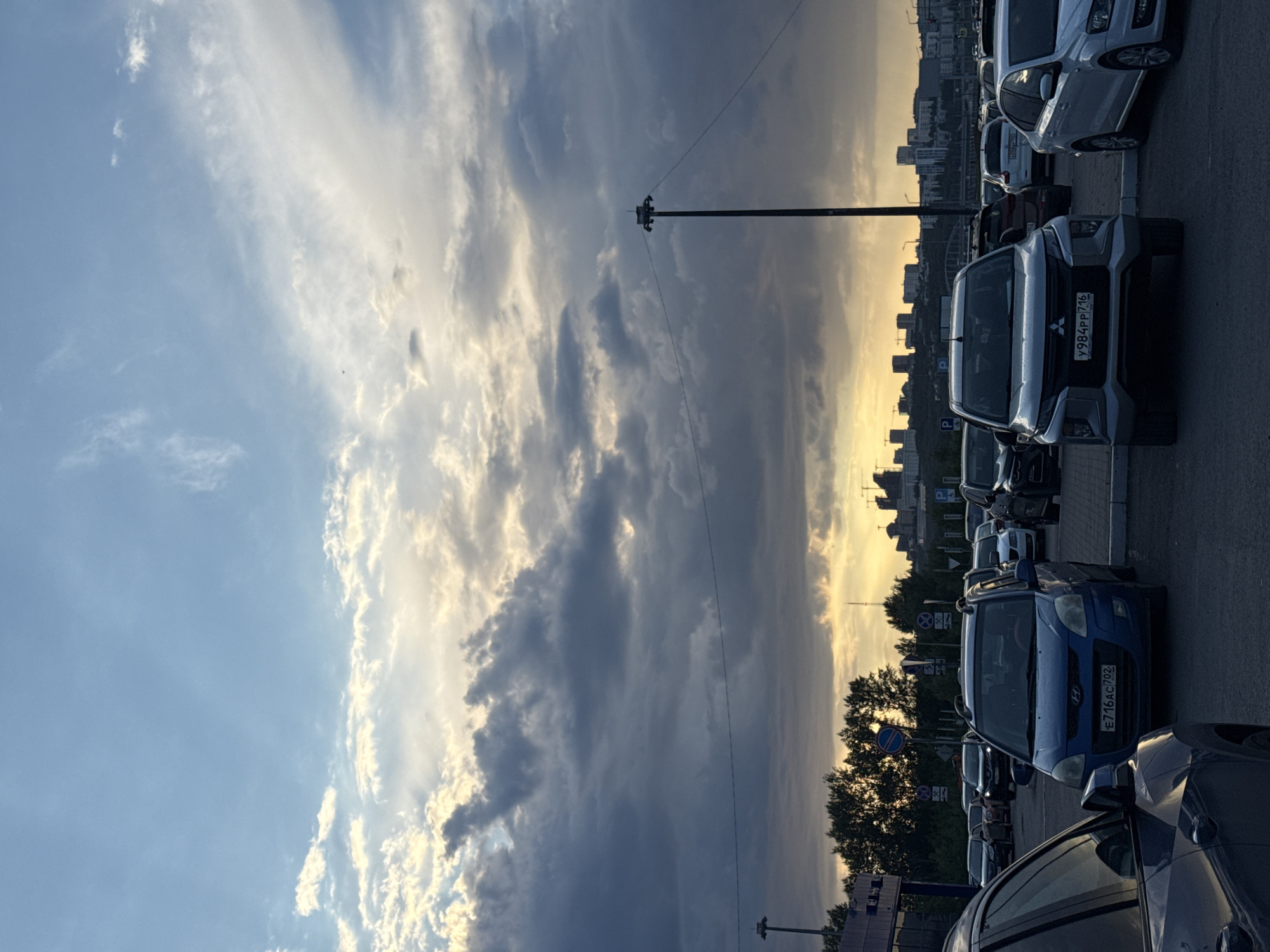 This is a low-angle, full shot of a parking lot with a city skyline and dramatic sky in the background. The sky is filled with heavy, dark clouds illuminated by streaks of sunlight, suggesting either dawn or dusk. Several cars are parked in the lot, including a blue Hyundai in the foreground, a silver Mitsubishi, and a white Volkswagen. The city skyline in the distance features numerous buildings and construction cranes. Trees line the edge of the parking lot. A tall light pole with wires is visible on the right side of the image.
