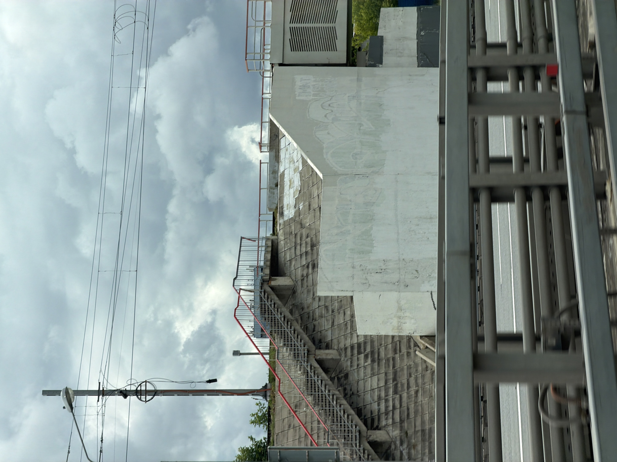 The image shows an outdoor scene in Moscow, Russia, under a cloudy sky. In the foreground, a grey metal guardrail is visible, with a highway or road behind it, blurred by motion. Beyond the highway, a concrete overpass structure dominates the midground. A wide, grey concrete staircase with red metal railings ascends the side of the overpass. The staircase is set against a textured, grey concrete embankment. At the top of the staircase is a walkway with railings and a large, white, industrial-looking structure with horizontal vents. The white concrete wall of the overpass is marked with white graffiti. To the left of the staircase, a tall utility pole with a lamp and electrical wires is visible, with green trees in the background. The overall impression is of an urban infrastructure setting.