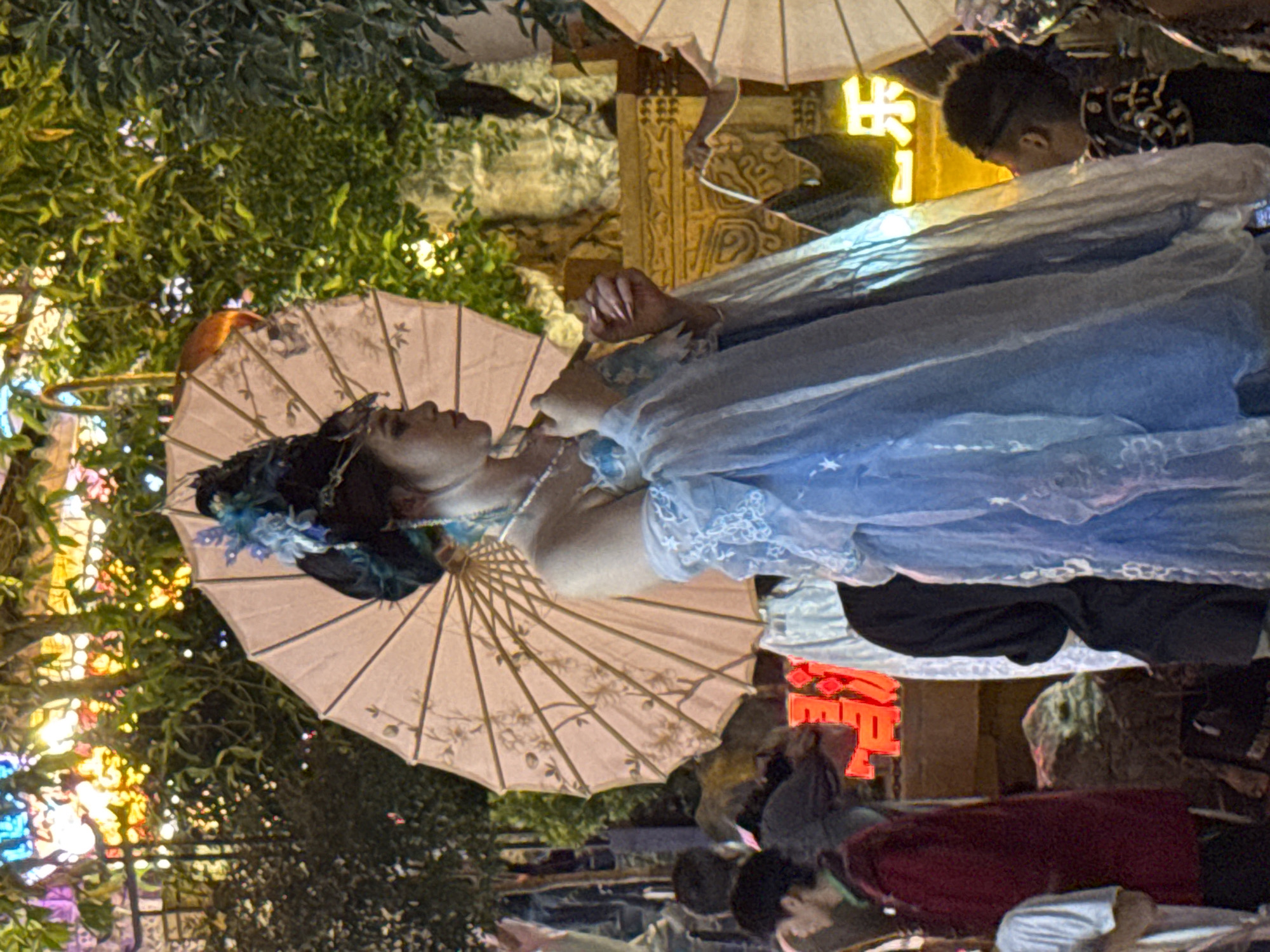 The image depicts a nighttime scene in Yuzhong District, China, featuring a woman in traditional Chinese attire holding a light pink umbrella. She is adorned with a blue and black headdress and a pearl necklace, and wears a blue dress with intricate white lace detailing. The background is illuminated by colorful lights and lush greenery, with other individuals visible in the periphery. The overall atmosphere suggests a festive or cultural event.