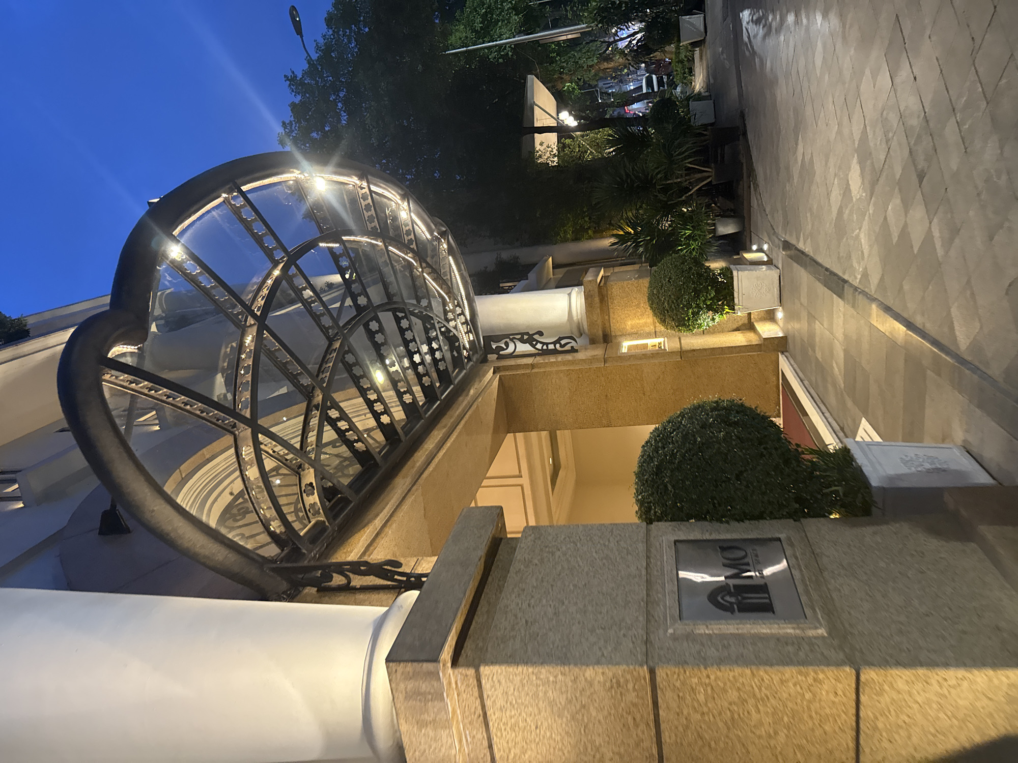 The image captures the entrance of the Mandarin Oriental Hotel in Hà Nội, Vietnam, at night. The entrance features a curved glass and wrought iron awning with integrated lighting. To the right of the entrance is a planter with a round bush and a sign displaying the "MO" logo. The facade is constructed from light-colored stone. A paved walkway extends into the distance. The sky is a deep blue, indicating dusk or night, and the surrounding trees are illuminated by streetlights.