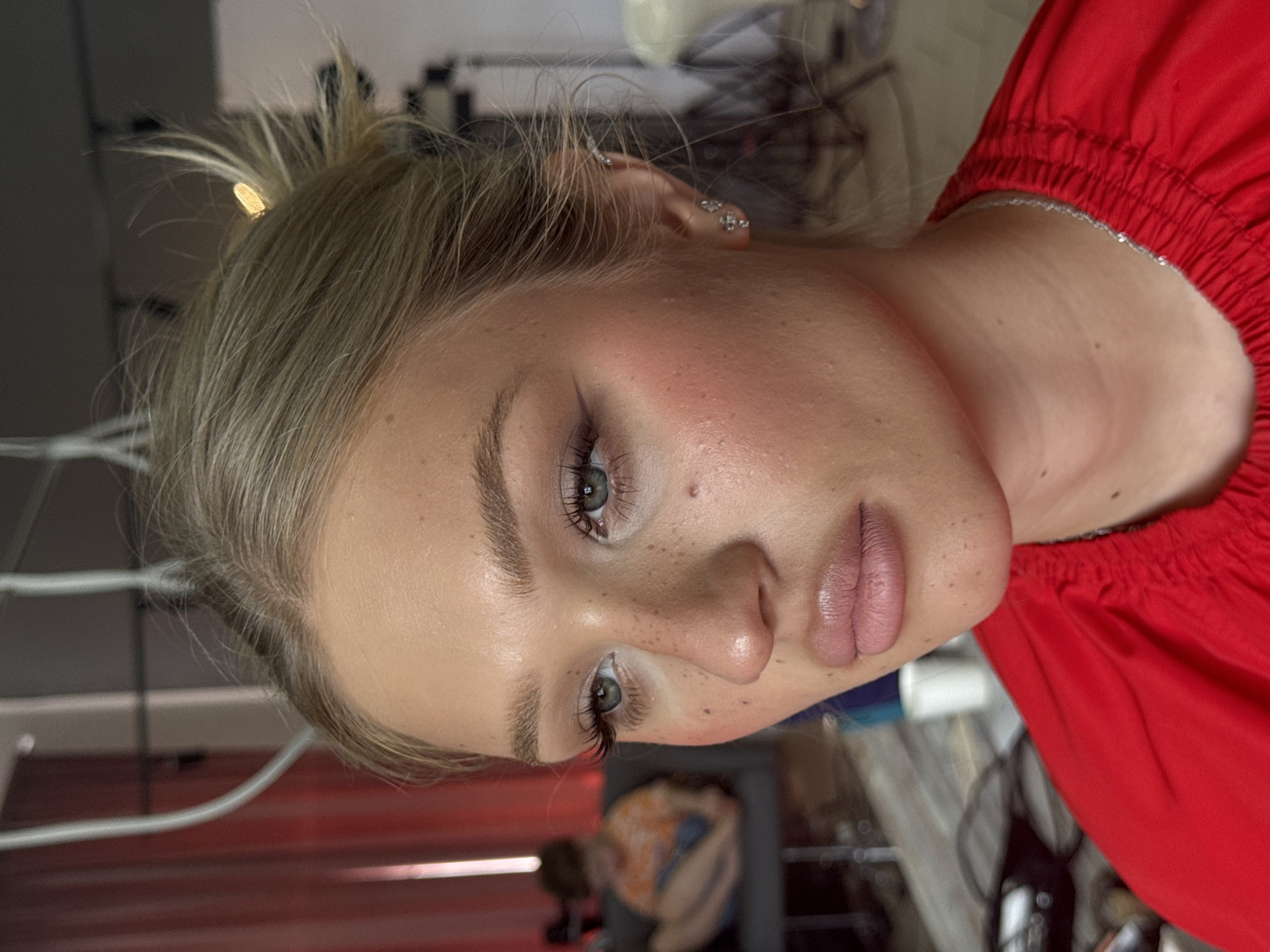 A close-up, side-profile, medium-shot captures a young Caucasian woman with blonde hair styled in a messy bun, a small golden hair clip visible. She is wearing a bright red garment and a silver necklace. Her makeup is artfully applied, featuring winged eyeliner, blush, and lip gloss, accentuating her freckled complexion. Two small, sparkling earrings adorn her left ear. In the blurred background, another person is seated on a couch, wearing an orange and white shirt. The setting appears to be an indoor space, possibly a studio or dressing room, with soft lighting and a hint of red fabric hanging in the background. The image is a professional portrait, focusing on the woman's facial features and makeup. There is no discernible information linking this image to Novocherkassk, Russia.