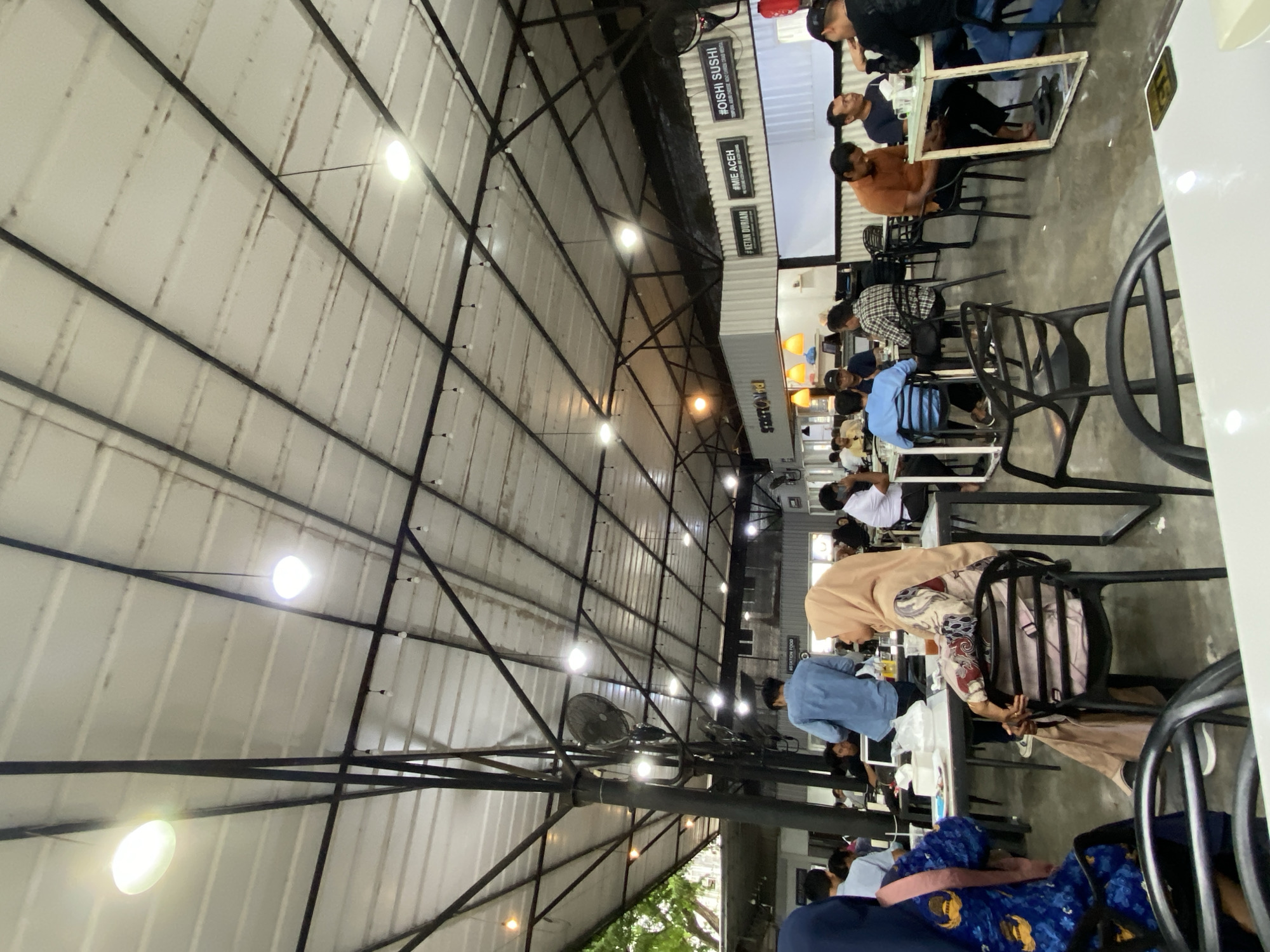 The image depicts a bustling, semi-outdoor dining area, likely a food court or an eatery in Lhokseumawe, Indonesia, illuminated by artificial lighting, suggesting it is evening or night. The space is covered by a high, pitched roof made of corrugated metal panels, supported by a framework of black metal trusses and beams from which numerous spherical white lights are suspended. Ceiling fans are also mounted on some of the support structures.
A crowd of people, mostly adults, are seated at various tables, predominantly black chairs, engaged in dining, conversation, or waiting for food. In the foreground, a woman wearing a beige hijab and a patterned blouse is seated at a table, with another person in a blue denim-like shirt visible nearby. Further back, many more patrons fill the seating areas.
To the right side of the image, several food stalls or vendor units are visible, constructed from white corrugated metal. These stalls feature prominent signs with hashtags indicating the cuisine they offer. Visible text includes "#OISHI SUSHI" with smaller text "REAL AND TASTY, NOT CHINESE," "#MIE ACEH" with "TRADITIONAL CUISINE OF ACEH" underneath, and "#KETAN DURIAN." Another sign reads "RESTORAN FOOD." The floor appears to be concrete, and some tree leaves are visible towards the lower left, indicating the open nature of the venue. The overall atmosphere appears casual and lively.