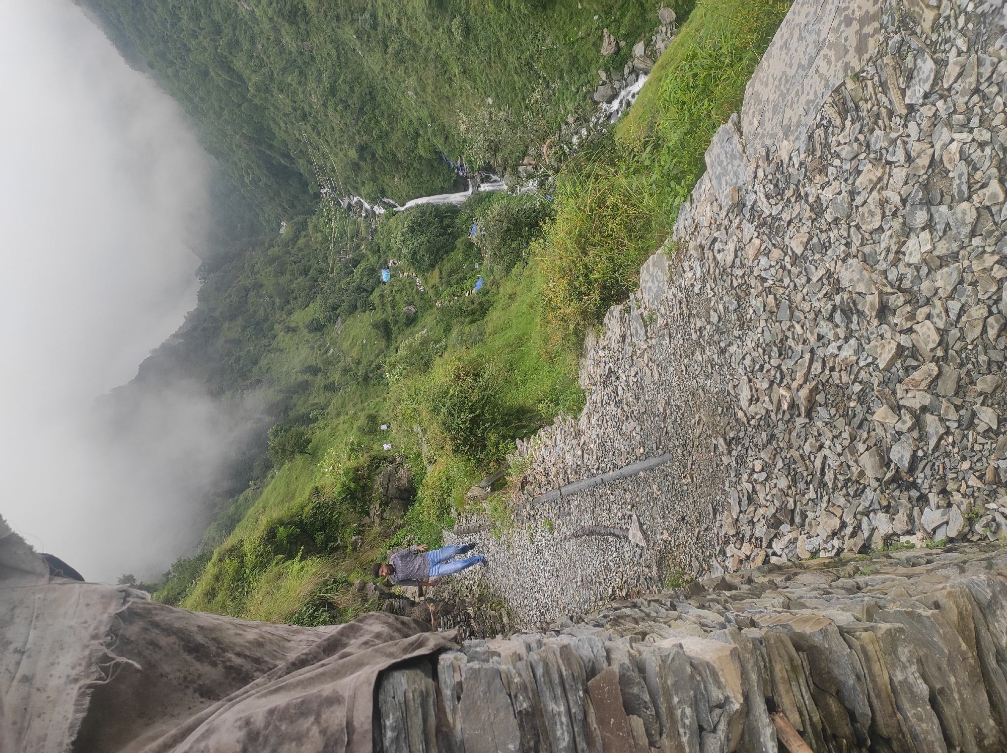 In Bhagsu, India, a man walks on a gravel path along a steep mountain slope covered in lush green vegetation. The path is bordered on one side by a stone wall and on the other by the incline of the mountain. In the background, a waterfall cascades down a rocky valley, shrouded in mist. Several blue tents are visible near the waterfall, suggesting a campsite or resting area. The sky is overcast, contributing to the misty atmosphere.