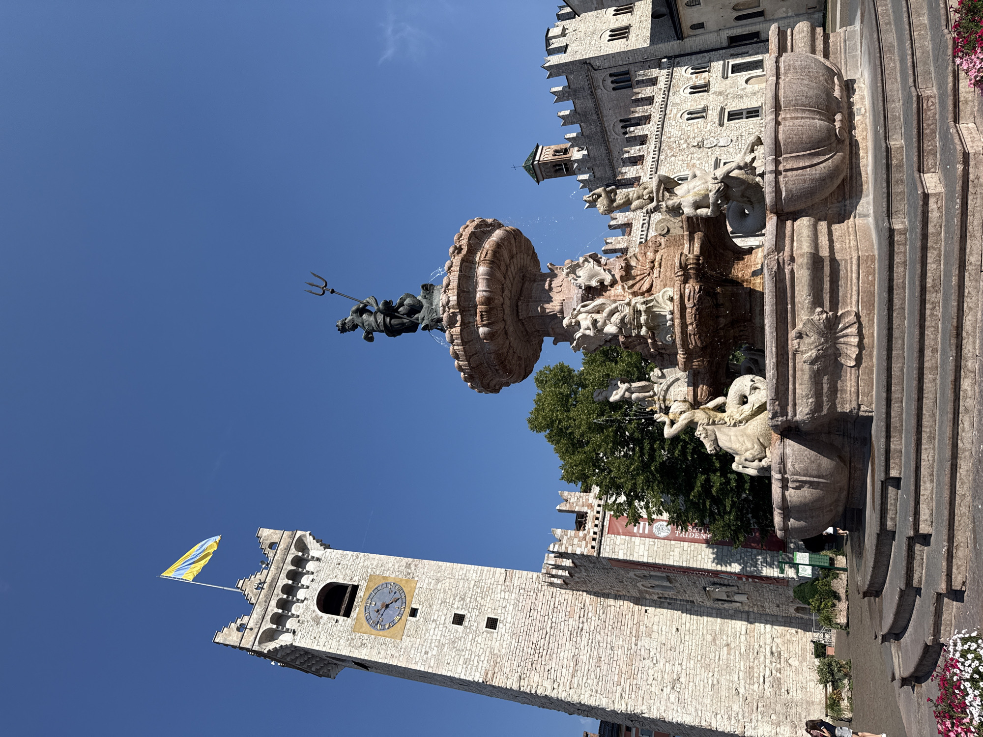 The image depicts Piazza Duomo in Trento, Italy, dominated by the Fontana del Nettuno and the Torre Civica. The Fontana del Nettuno, a large Baroque fountain, is in the foreground, featuring multiple tiers adorned with sculptures of mythological figures, including Neptune at the apex holding a trident. Water streams from various spouts, creating a dynamic effect. To the left, the Torre Civica, a tall, square clock tower constructed of white stone, rises against a clear blue sky. A flag, predominantly yellow and blue, flies from its summit. The tower has a large, circular clock face with Roman numerals. Behind the fountain and tower, a long, imposing building with arched windows and crenellated battlements, likely part of the Palazzo Pretorio or Palazzo della Loggia, stretches across the mid-ground. The sky is a bright, cloudless blue, indicating a sunny day. A few people are visible in the scene: one person in the lower left foreground near some flowers, and two individuals in the middle ground to the left of the tower, appearing to walk or stand. Lush green trees are visible behind the fountain, providing a contrast to the stone architecture. The overall scene is one of historical significance and public space, bathed in bright sunlight.