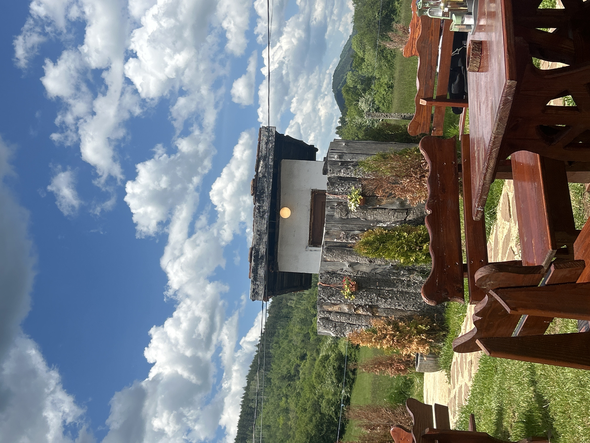 The image depicts a scene in Meshtitsa, Bulgaria. Dominating the view is a rustic wooden building with a white second story and a dark, possibly tiled roof. A light fixture is visible on the side of the building. In the foreground, there is a wooden table and chairs, likely part of an outdoor seating area. The environment includes a green, grassy area with various plants and shrubs. In the background, there is a wooded hillside under a bright blue sky with scattered white clouds. Powerlines stretch across the sky, running diagonally from top to bottom.