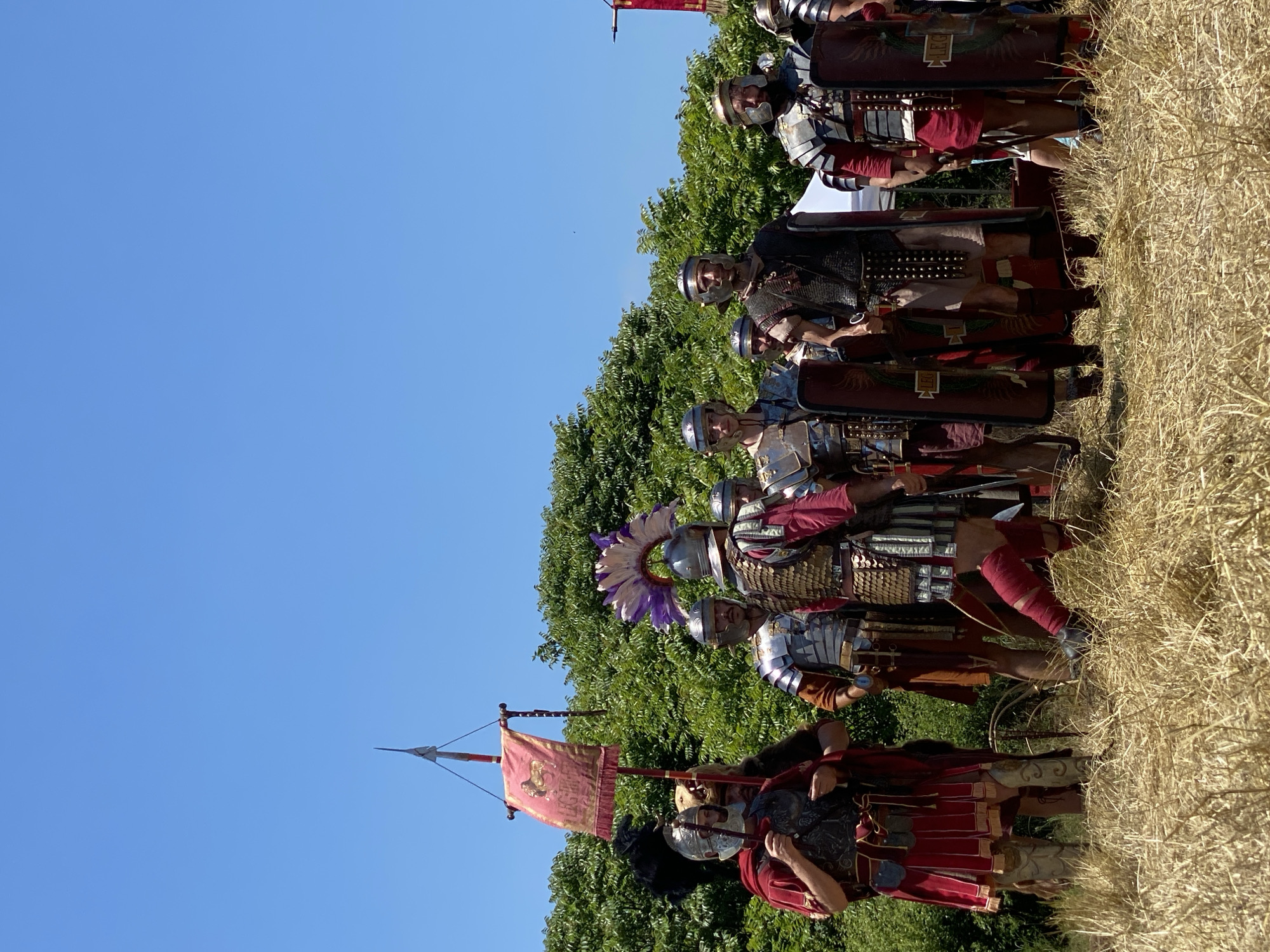 The image depicts a group of approximately ten individuals dressed as Roman soldiers, participating in an outdoor historical reenactment in Gigen, Bulgaria. The scene is set on a field of dry, light-brown grass under a clear, bright blue sky. A dense line of green deciduous trees and bushes forms the background.
In the foreground, the Roman reenactors are arranged in a staggered line. From left to right:
1. A figure in a red tunic, red cloak, and silver helmet, holding an object, stands next to a red vexillum (standard).
2. Behind this figure, another individual wears a helmet adorned with a bearskin.
3. The vexillum, a rectangular red banner with golden trim and tassels, is suspended from a vertical pole topped with a spearhead. A golden lion emblem and the inscription "LEG" are visible on the banner.
4. Further right, a reenactor wears a silver helmet with a prominent purple and white feathered plume. They are dressed in lorica segmentata (segmented plate armor) over a brown tunic.
5. Other participants wear various forms of Roman armor including lorica segmentata, lorica squamata (scale armor), and possibly lorica hamata (chainmail), over tunics in shades of red, brown, and dark colors.
6. Several rectangular shields (scuta), predominantly dark red/brown, are visible, leaning against the soldiers' legs. Some shields display the inscription "LEG".
7. The individuals are standing upright, facing generally towards the viewer's right, appearing to be assembled or awaiting instruction.
8. Footwear consists of boots or sandals, with some individuals wearing red leg wraps.
The overall impression is a static formation of historically costumed figures in a rural outdoor setting.