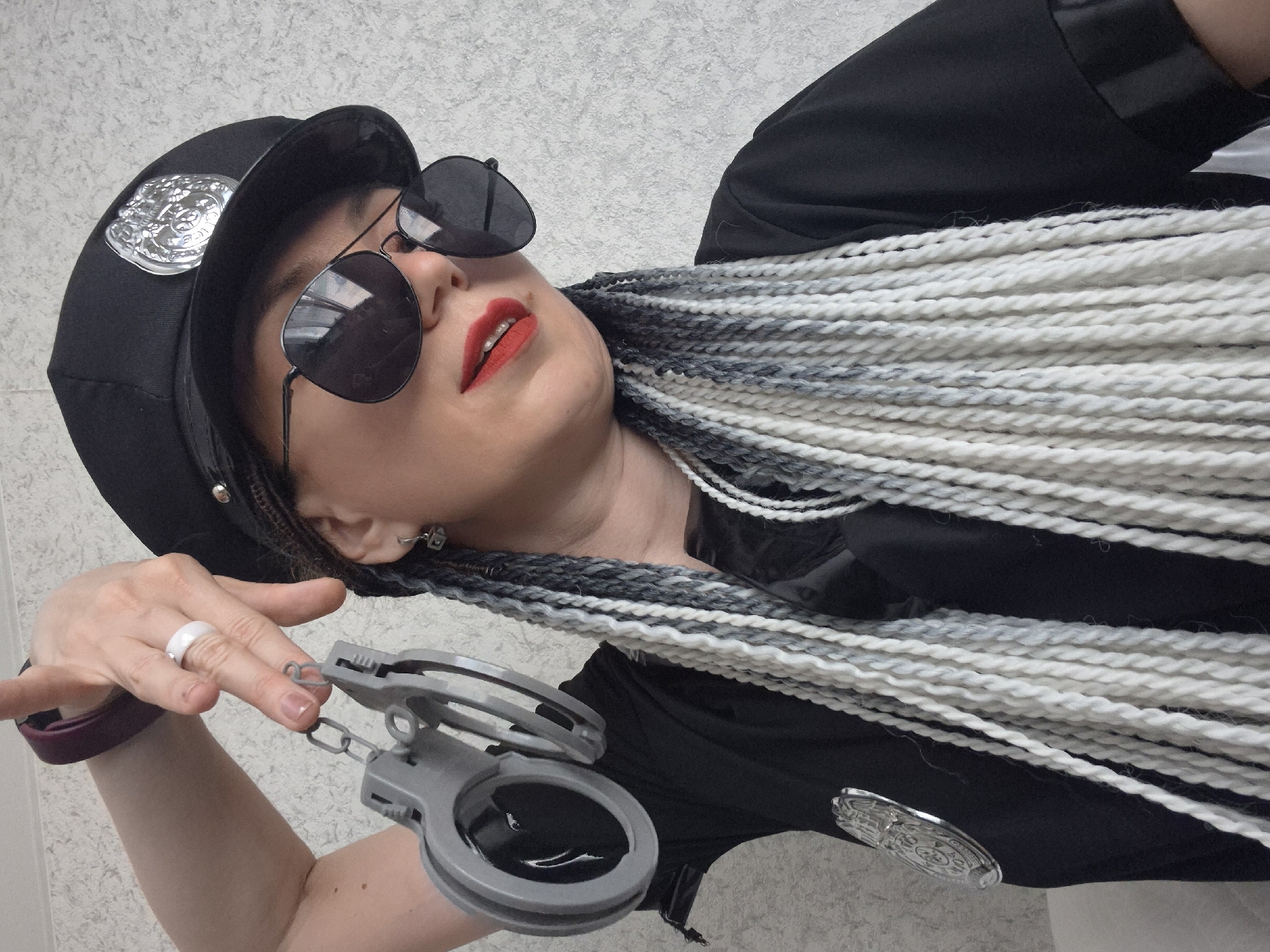 A person wearing a police hat and sunglasses holds a pair of handcuffs. They have braided hair with white and grey streaks. The person is wearing a black uniform shirt with a badge on the left side. The background appears to be a textured wall. There is no information available about the location in the city of Chaikovsky, Russia.