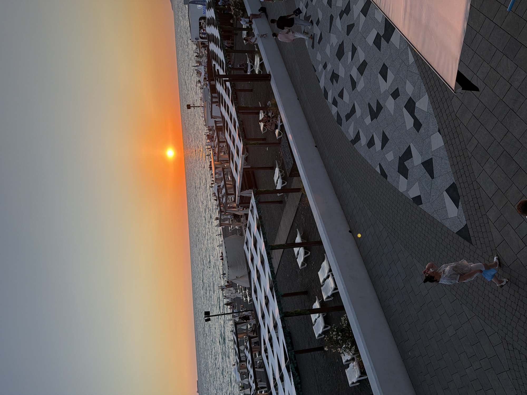 This image shows a sunset view of a beach in Sochi, Russia. The sun is low on the horizon, casting an orange glow over the ocean. The beach is lined with lounge chairs and umbrellas, and there are several people walking along the shore. In the foreground, a woman is walking on a paved walkway. The scene is peaceful and serene, capturing the beauty of a sunset at the beach.