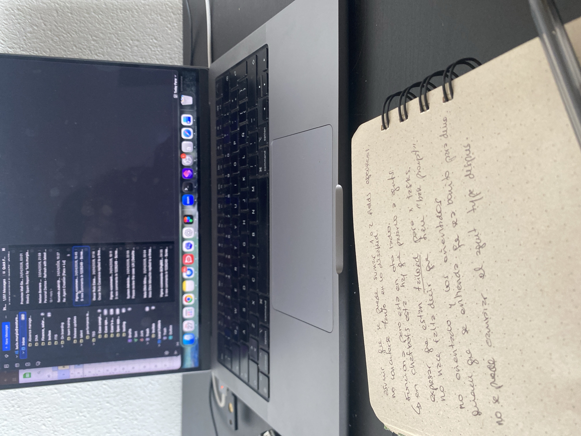 The image shows a close-up of a workspace. A silver Apple MacBook Pro is partially open, displaying an email inbox on the screen. To the right of the laptop is a small notebook with spiral binding. Handwritten notes in Spanish fill the visible page of the notebook. A pen rests along the edge of the notebook. The desk appears to be black or dark gray, with a white wall in the background.