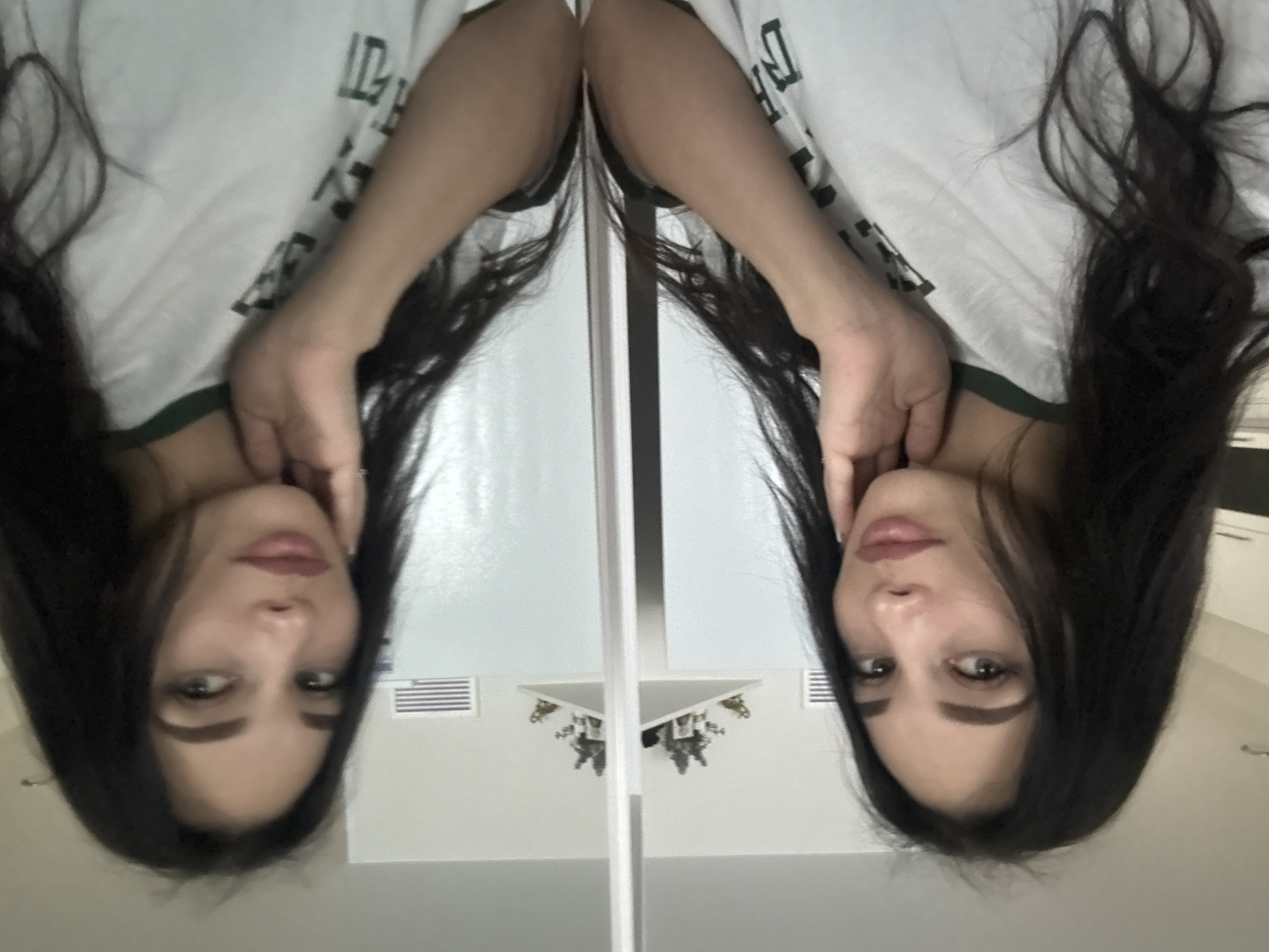 A woman is shown reflected in a mirror. She has long, dark hair, fair skin, and is wearing a white t-shirt with dark green trim. The mirror's reflection shows a white ceiling with a light fixture, an air vent, and what appears to be a cabinet in the background. It is not possible to confirm the location is Chaikovsky, Russia from the image provided.