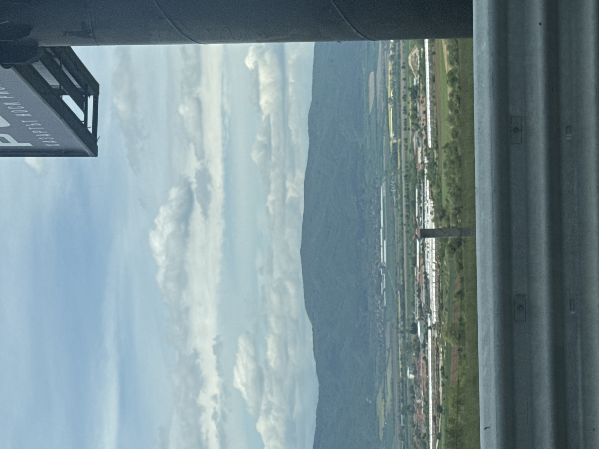 The image shows a view from an elevated position, likely a building or structure. The foreground includes parts of the structure itself: a grey corrugated metal column on the right and what appears to be a pipe or beam overhead. On the left, there's a partial view of a sign with text "BAPTIST HOCK PRO". Beyond the structure, the landscape reveals a mountain range in the middle ground, with a sky featuring light blue patches and scattered white clouds. A linear arrangement of buildings or structures is visible along the base of the mountain, interspersed with green fields or vegetation. The scene is located in Unknown, Unknown.