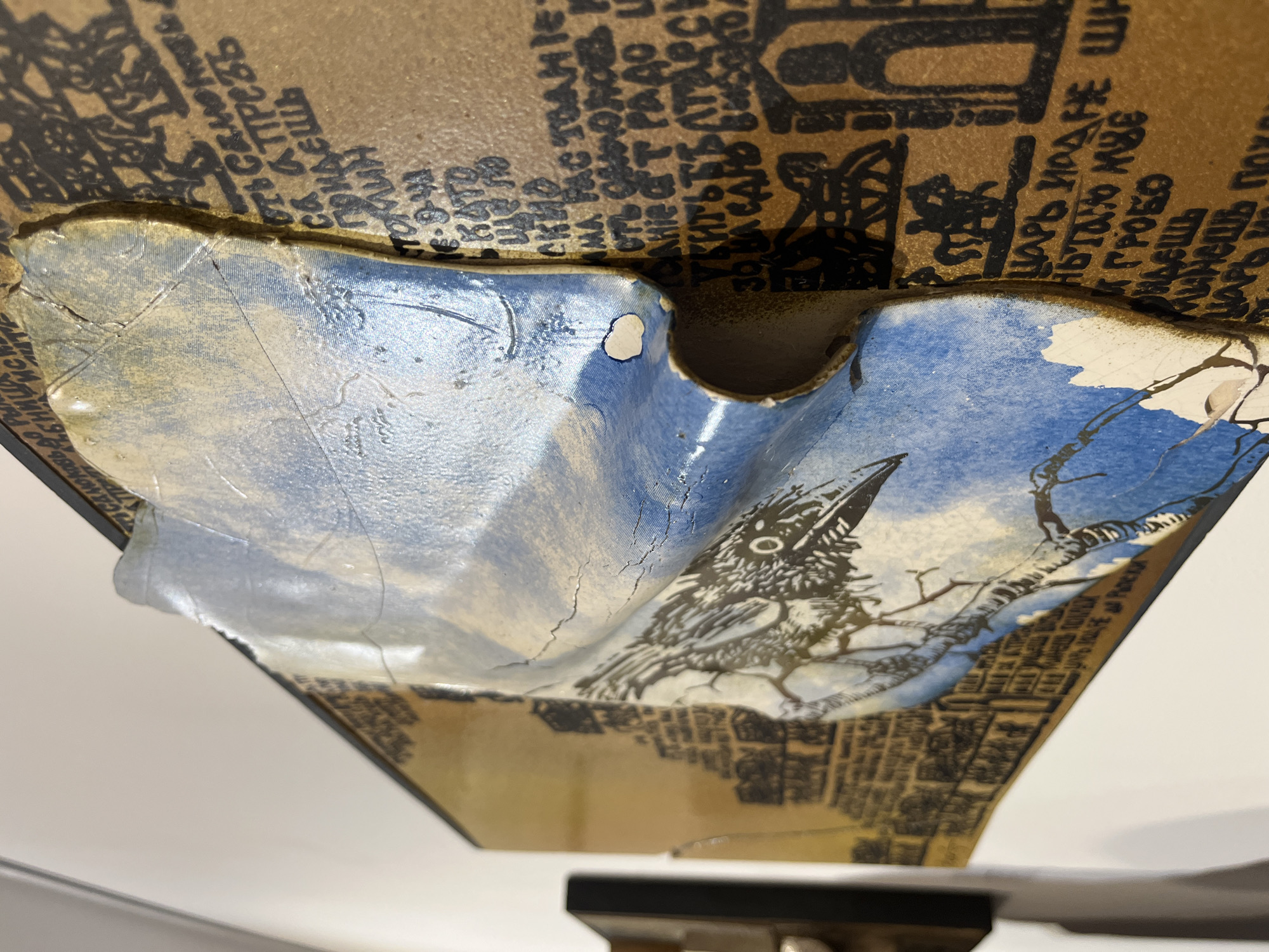 This media file displays a close-up of a complex, multi-layered art piece, likely a decorative panel or a segment of a larger installation, viewed from an oblique, upward-looking angle. The object exhibits a distinct curved, sculptural quality.
The uppermost layer is a flat, brownish-gold surface extensively covered with black Cyrillic text and faint, abstract illustrations, giving it the appearance of aged parchment or printed material. Below this, a prominent, concave segment forms a central element. This segment is painted with a sky-blue background, featuring wispy white clouds, and prominently depicts a stylized black and white crow or raven perched on a bare tree branch. The painted surface of this blue section shows notable cracking and distress, suggesting age or a deliberate artistic effect.
Beneath the curved crow motif, another flat, brownish-gold layer emerges, also adorned with black Cyrillic script and intricate patterns that resemble a cityscape or abstract architectural forms. A dark, structural element is visible at the very bottom edge, suggesting the art piece is mounted or part of a larger structure.
Visible Cyrillic text, partially fragmented across the layers, includes words such as "ПОЙДЕШЬ" (you will go), "СА СТРЕСС" (with stress), "КАК" (as/how), "ЦАРЬ ИОДА НЕ ШИ" (Tsar Iod does not sew/stitch), "ГРОБО" (coffin/grave), "ПАРК" (park), and "ПЛОХА" (bad). These textual elements, combined with the imagery of the crow and cityscape, contribute to a somber, historical, or reflective artistic narrative. The use of Cyrillic text is consistent with the provided location context of Plovdiv, Bulgaria. The overall impression is one of a richly textured and symbolically charged mixed-media artwork.