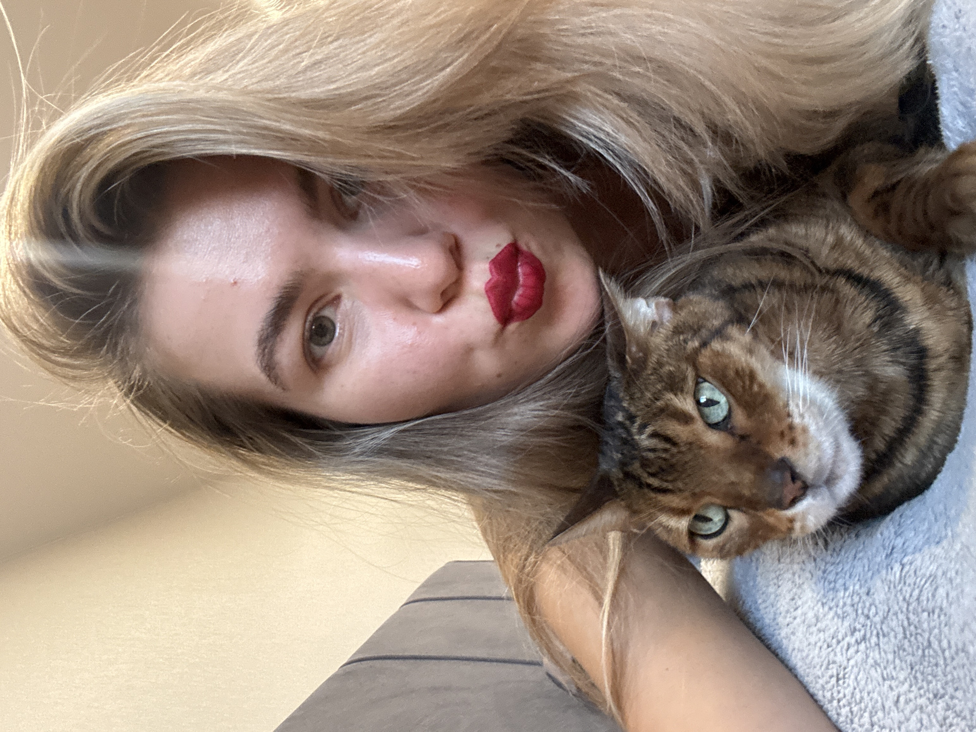 A selfie-style image presents a fair-skinned woman with long, blonde hair and striking red lipstick, juxtaposed with a brown tabby cat. The cat's face is close to the camera, displaying green eyes and a white muzzle. A grey textured surface is visible beneath the cat. The setting appears to be indoors with a pale yellow wall and a glimpse of a grey, striped surface in the background. Location: Unknown, Unknown.