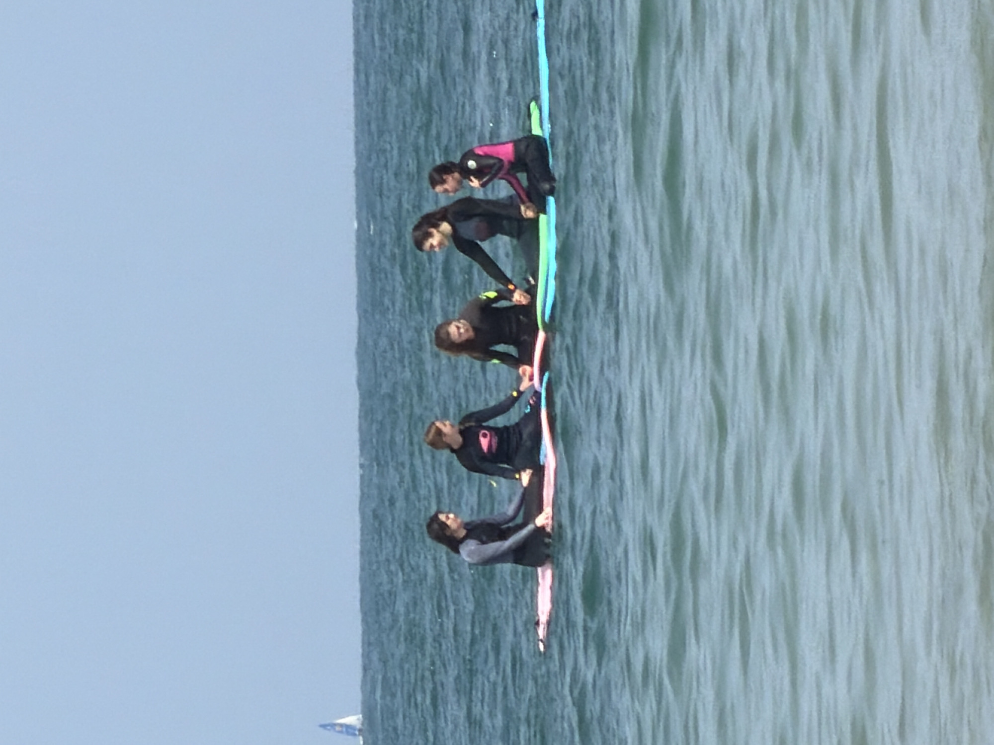 Five individuals wearing wetsuits are positioned on a long surfboard in the sea. They appear to be in a line formation. The sea has small ripples on the surface. The sky is light blue and clear. A boat sail is visible in the distance. Based on the image content, it could be in a coastal area like Tel-Aviv, Israel.