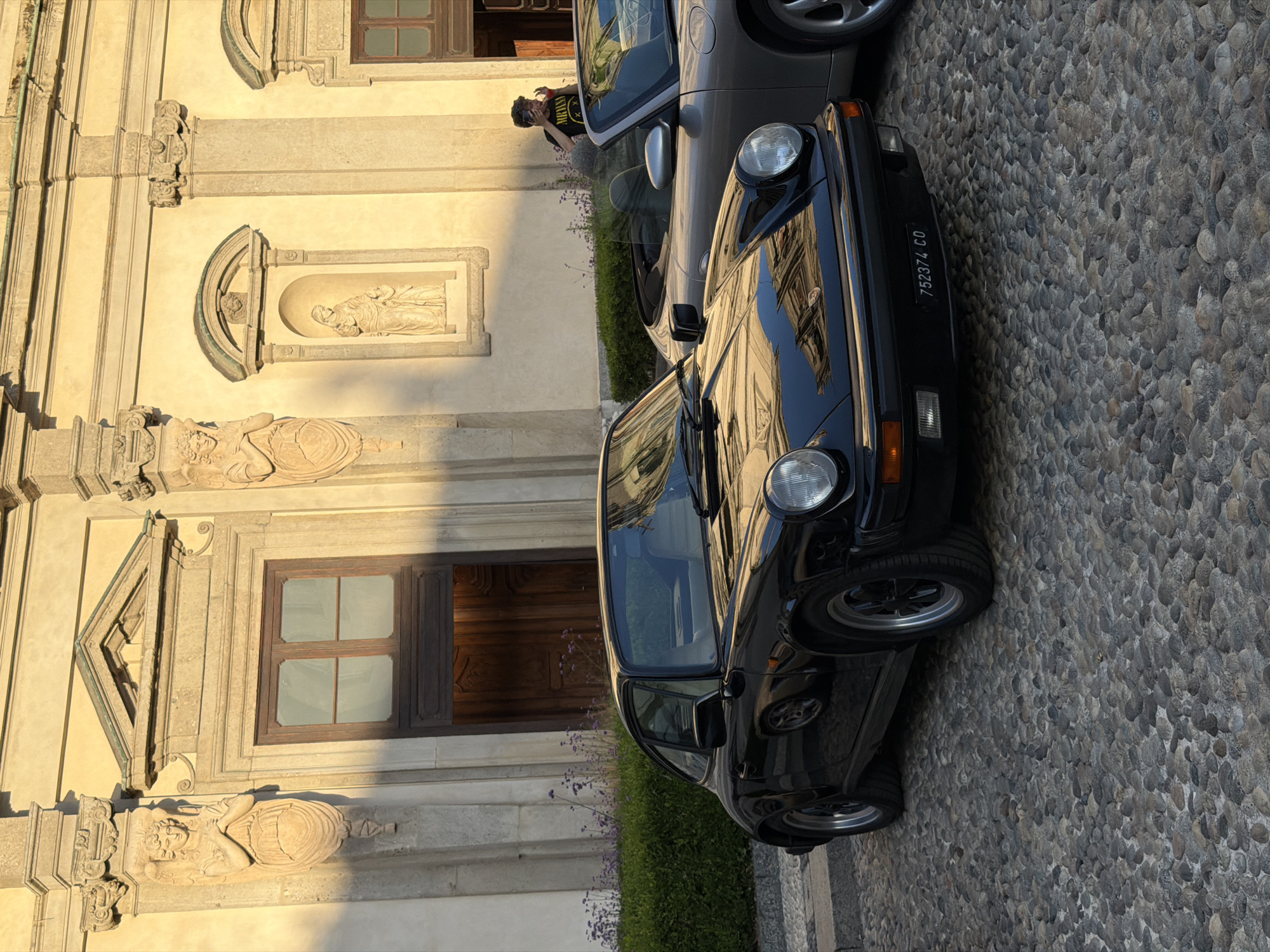 A black Porsche 911 coupe and a silver convertible Porsche are parked on a cobblestone street in front of a building with ornate architectural features. The building appears to be a church or historic structure, with detailed stone carvings of figures and religious imagery. The sunlight casts strong shadows across the facade and the cars. A person wearing a black t-shirt with a "Nirvana" logo is visible in the background, holding up a phone, possibly taking a photo. The scene is set in Milan, Italy, with a license plate on the black Porsche reading "752374 CO".