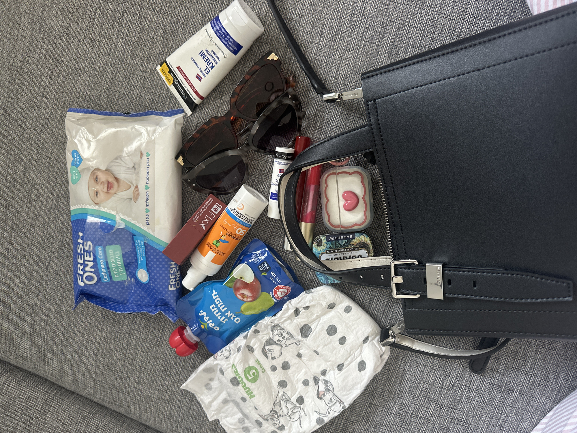 A black leather purse sits on a grey fabric sofa. The purse is open, revealing several items inside and around it. These items include a Huggies diaper with a Dalmatian print, a blue pouch of baby food with an apple and pear illustration, a tube of Neutrogena El Kremic sunscreen, a tube of Roche-Posay sunscreen for children, a red lipstick, a small heart-shaped case, two pairs of sunglasses, and a packet of Fresh Ones Cashmere Care baby wipes. The overall scene suggests a person is packing or unpacking a bag, possibly for a child. The image was taken in Petah Tikva, Israel.