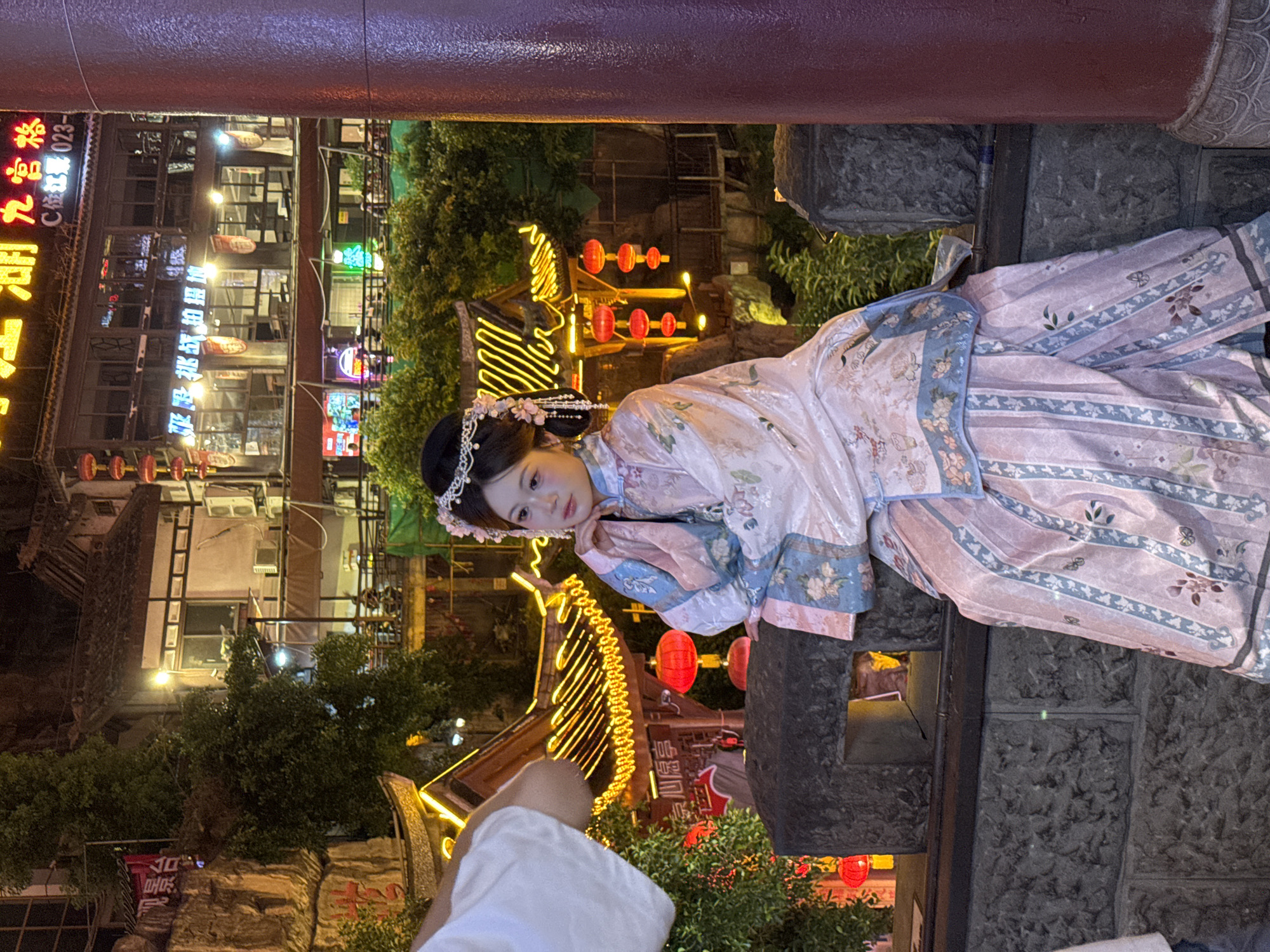 A young woman in a pink and blue traditional Chinese dress sits on a stone ledge at night. She has her hand on her chin and is looking to the left. The background shows a street with traditional architecture, illuminated by neon signs and lanterns. The location appears to be the Ciqikou Ancient Town in Yuzhong District, Chongqing, China, known for its historic buildings and vibrant nightlife. The woman is adorned with a floral hairpiece. The scene is lit by artificial lights, creating a warm and inviting atmosphere.