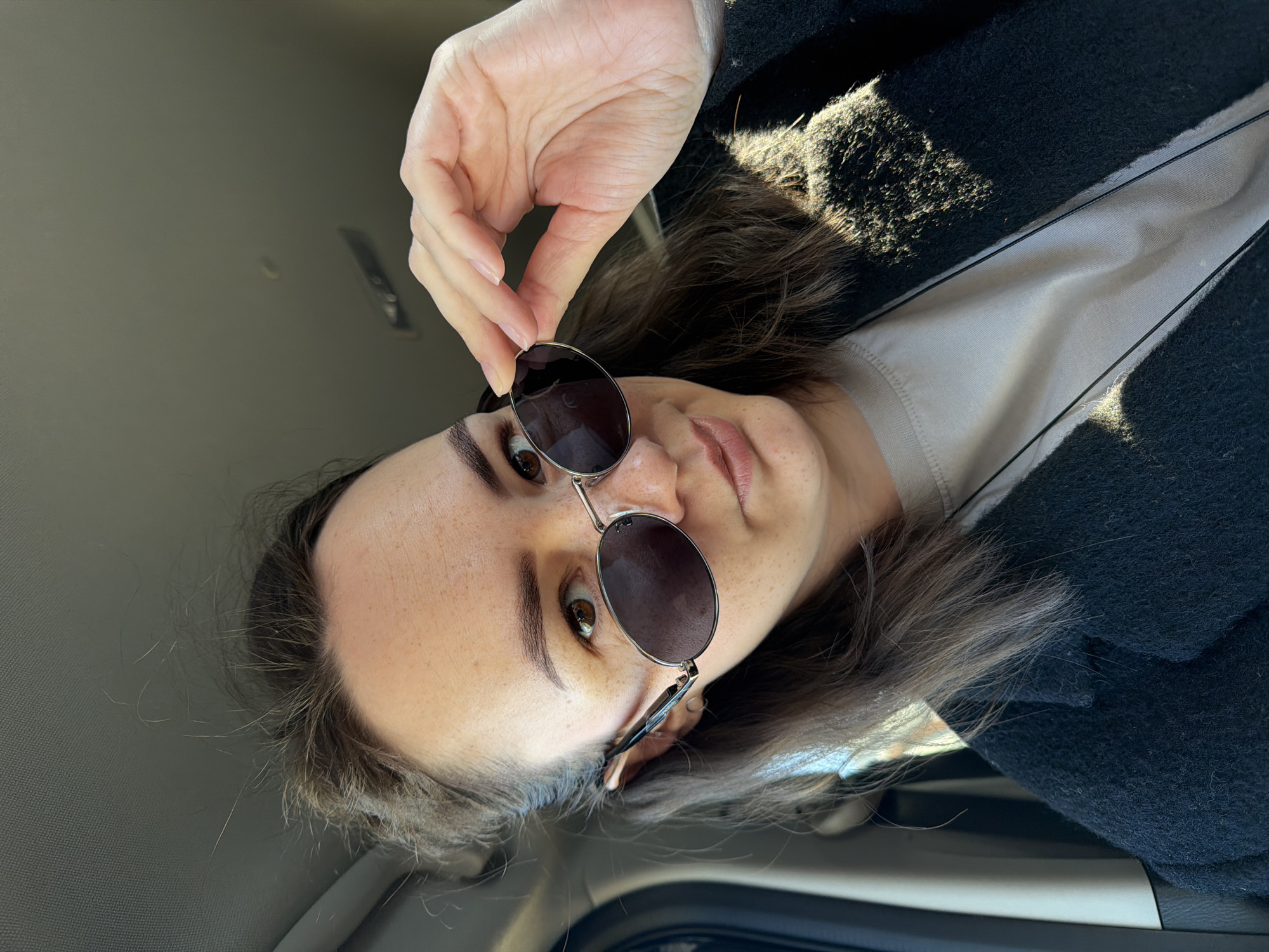 The rotated image captures a woman from a slightly low angle, positioned inside what appears to be a vehicle. She has medium brown hair, brown eyes, and visible freckles on her face. She is wearing a dark, textured jacket or coat over a lighter-colored shirt. Her hand, with neatly trimmed nails, holds a pair of round, metallic-framed sunglasses with dark lenses, resting just below her eyes. She looks directly at the camera with a neutral expression.
Bright sunlight streams in, casting strong highlights on her hair, the textured fabric of her jacket, and parts of the car's interior. The background shows the grey headliner or roof of the vehicle. Faint text, "FU," is visible on the right lens of her sunglasses. The setting is indoors, likely within a car, during daytime with clear, sunny weather. The location context is Chaikovsky, Russia.