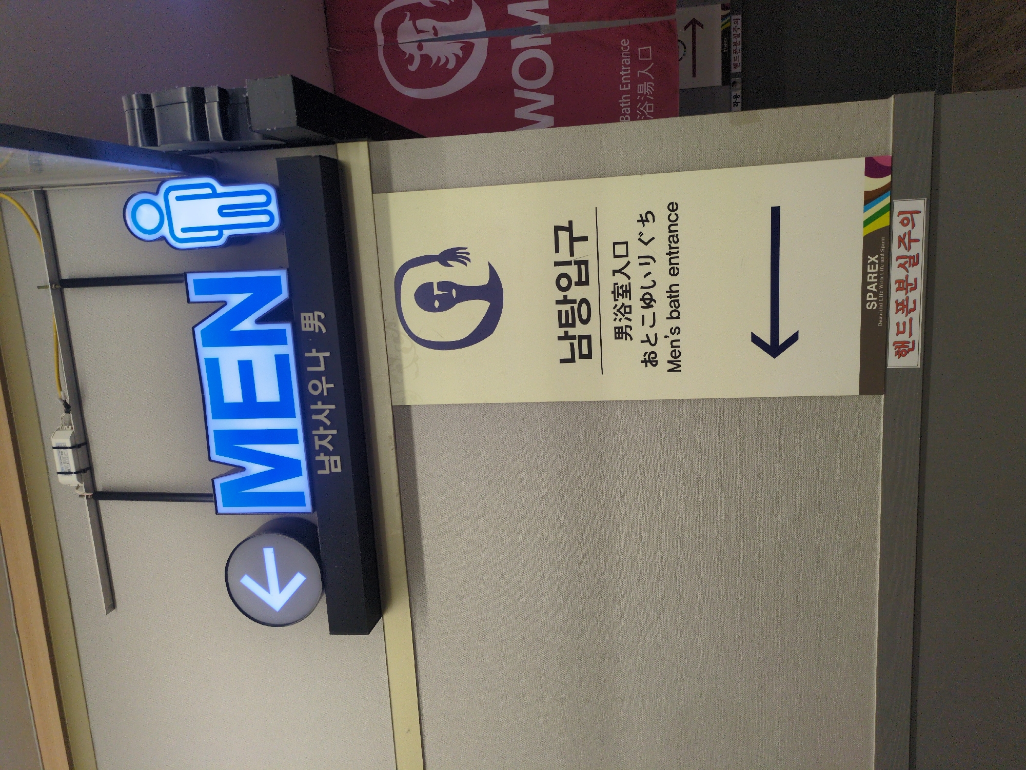 The image depicts signs indicating the location of a men's bath entrance in a commercial setting, possibly a spa or bathhouse. The signs are written in multiple languages, including English, Korean, and Japanese. The main sign features the word "MEN" in illuminated blue letters above a downward-pointing arrow. Adjacent to it is another sign with a stylized image of a man and text indicating "Men's bath entrance" in the three languages. A sign above references women's bath entrance. A safety warning concerning cell phone loss is also present. The presence of Korean text suggests the location is likely in South Korea, specifically in Seoul, if considering the commonality of multilingual signage in tourist areas.