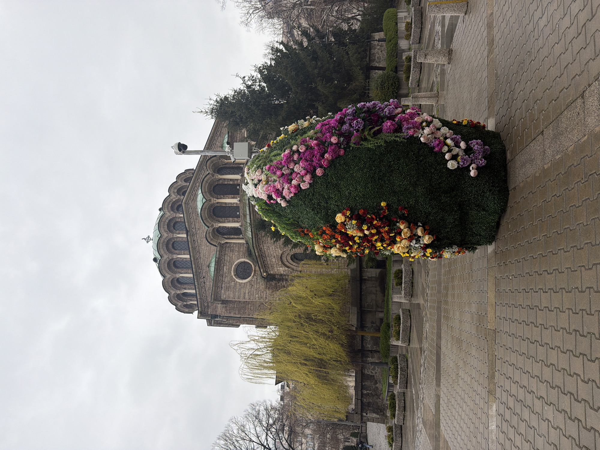 The image shows the St. Alexander Nevsky Cathedral in Sofia, Bulgaria. A large, egg-shaped floral arrangement is prominently displayed on the right side of the frame. The cathedral's facade is visible in the background, showcasing its architectural design, domes, and windows. The sky is overcast, suggesting a cloudy day. A paved walkway or plaza is in the foreground, with a pattern of interlocking stones. There are also small plants and possibly trees in the surrounding areas, adding to the overall environment.