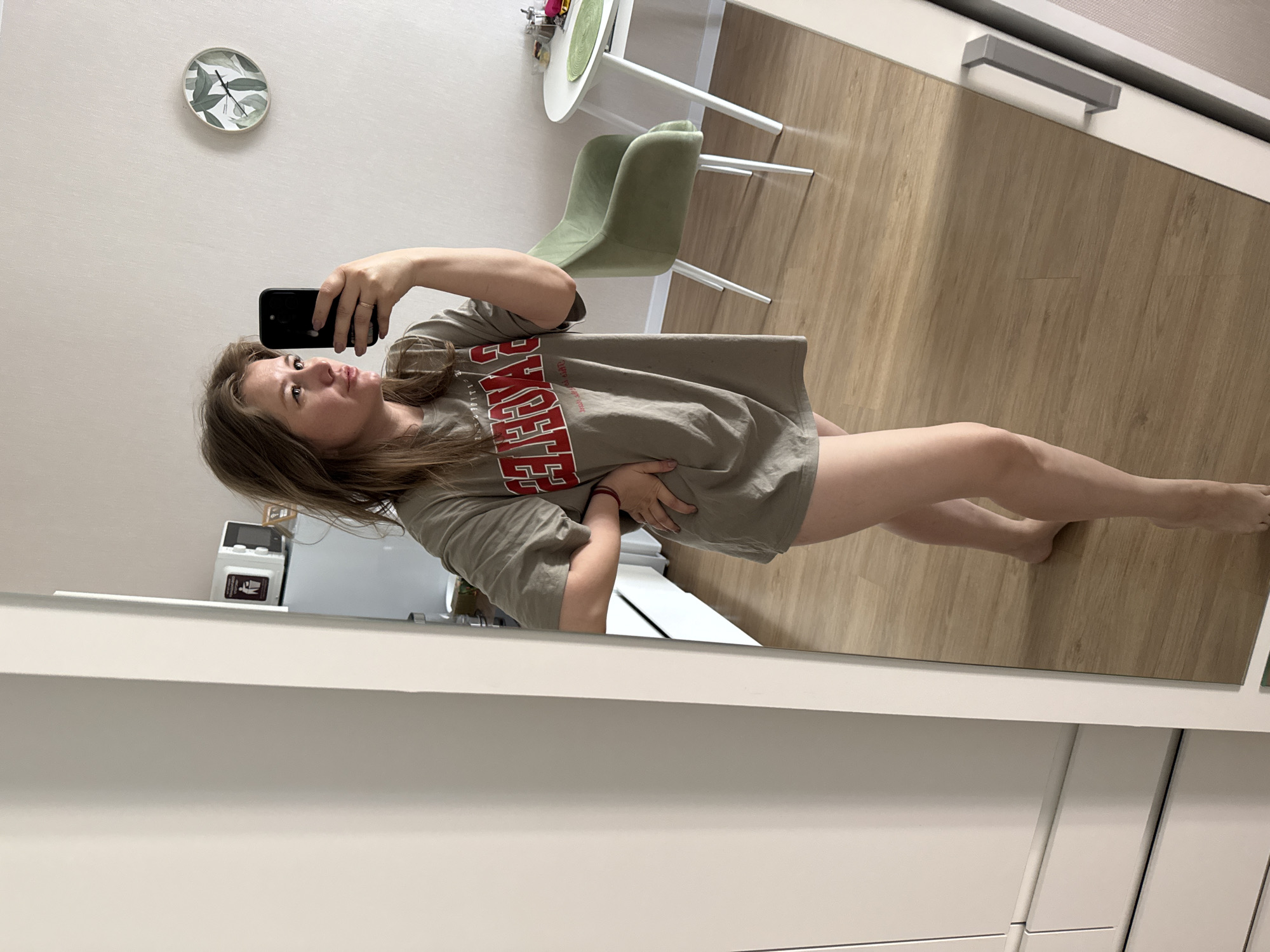 A young woman is captured in a mirror selfie, holding a black smartphone to take the picture. She has light brown hair and is wearing a loose-fitting, light brown/tan t-shirt with the word "EAGLES" prominently displayed in red, block letters with a white outline across the chest. Her left hand is placed on her stomach, and her legs are visible, suggesting the t-shirt is worn as a dress or with very short shorts.
The setting appears to be an indoor room, likely a modern home. The background features light-colored walls and a light wood laminate floor. On the wall, a round clock with a subtle green leaf design is visible. To the left of the woman, a small, round white table with white legs and a light green upholstered chair with matching white legs are present, suggesting a dining or casual seating area. Further in the background, a white cabinet or piece of furniture with a silver handle is visible. In the reflection near the bottom, white cabinetry or built-ins are also present. An appliance, possibly a small water dispenser or air purifier, with a small screen and a red symbol, can be seen behind her shoulder.
The woman looks directly towards her phone's camera, with a calm and slightly posed expression. The lighting is bright and even, indicating daytime. There are no specific visual cues within the image to confirm the provided location context of Perm, Russia.
