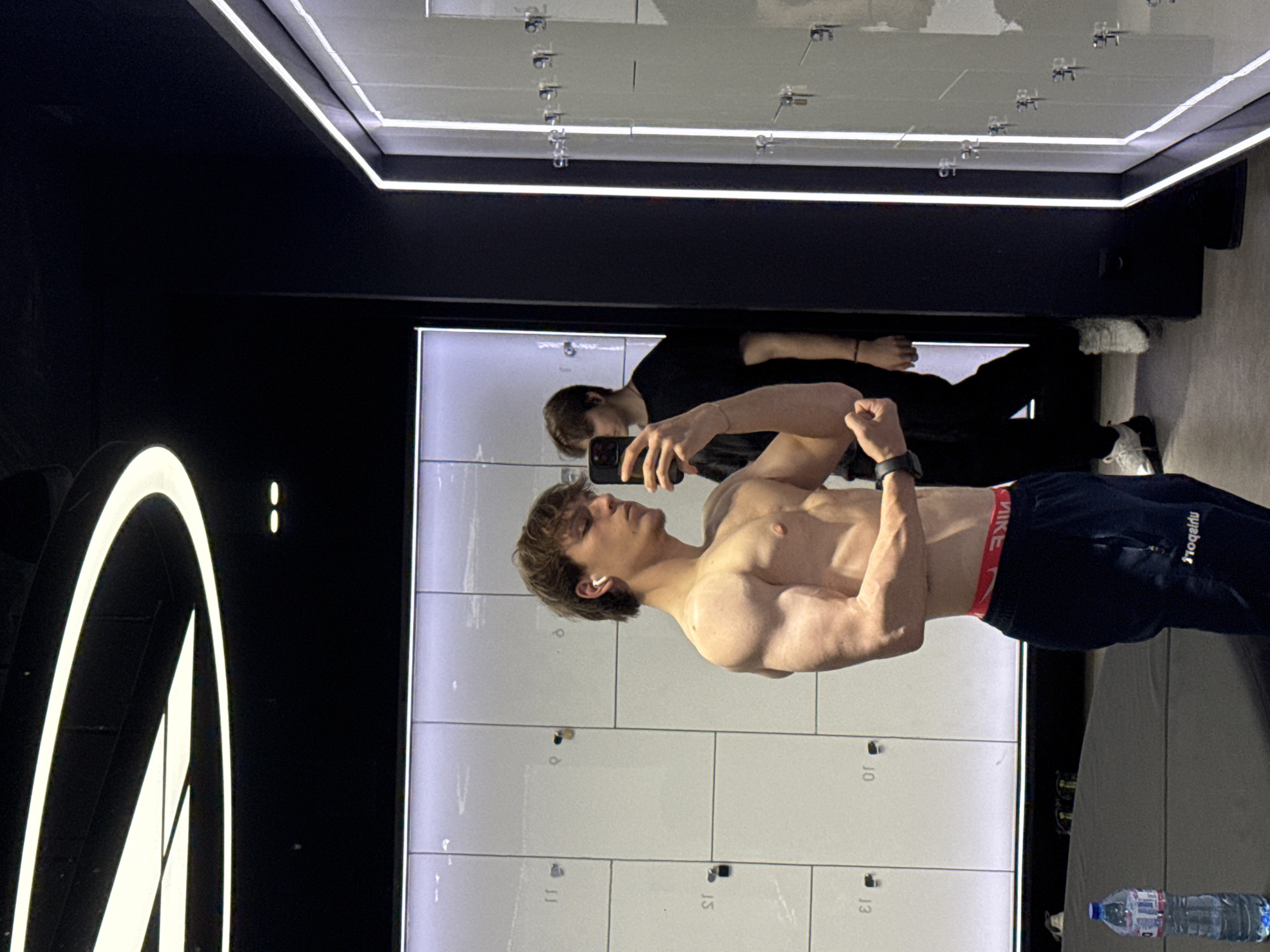 A young man, shirtless and with defined musculature, is captured in a mirror selfie in what appears to be a modern gym or locker room in Sofia, Bulgaria. He is seen from the waist up, flexing his biceps and looking intently at his reflection, holding a black smartphone to take the picture. He has short, light brown hair and is wearing white wireless earbuds. A black watch is visible on his left wrist. His athletic attire includes dark blue sweatpants with "Prozis" text visible on the left leg, and red "NIKE" branded underwear peaking above the waistband.
In the background reflection, another person wearing a black t-shirt is partially visible, also looking towards the mirror. The setting features rows of light-colored lockers, with numbers "10", "11", "13", and "15" discernible on their surfaces. The room has a contemporary design with prominent linear LED lighting integrated into the ceiling and surrounding the mirror. The floor is a dark gray, and a clear plastic water bottle with a blue cap is visible on the floor in the foreground. The overall ambiance is clean and well-lit, typical of a fitness facility.