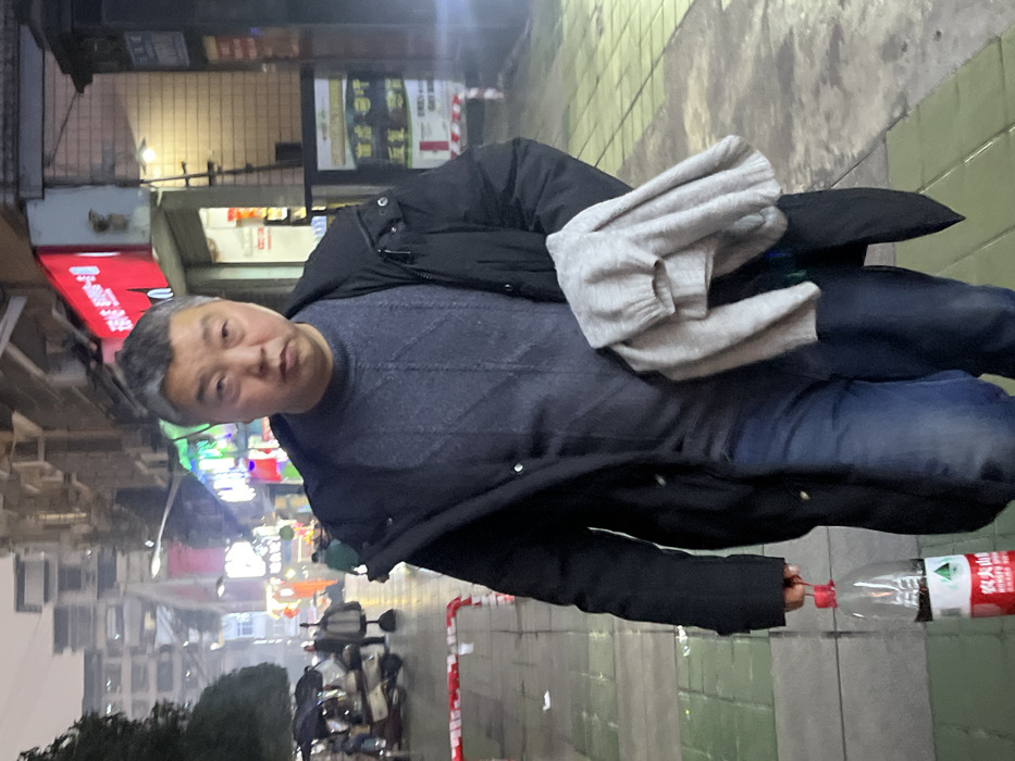 A male subject stands on a wet, paved sidewalk in an urban area identified as Jiangjin, China, during what appears to be evening or night. He is an East Asian adult, middle-aged, with short, greying hair and a neutral expression, looking forward. He wears a dark blue patterned sweater, dark blue jeans, and an open black long coat. In his left hand, he holds a clear plastic Nongfu Spring bottle containing a dark liquid. A light grey, folded sweater is carried over his right arm. The background to his left features additional blurred individuals, some holding umbrellas, parked motorbikes/scooters, and a red and white striped barrier on the sidewalk. To his right, illuminated storefronts with Chinese characters on red and green signs are visible along the street, backed by multi-story buildings. Artificial lighting from streetlights and shop signs illuminates the scene.