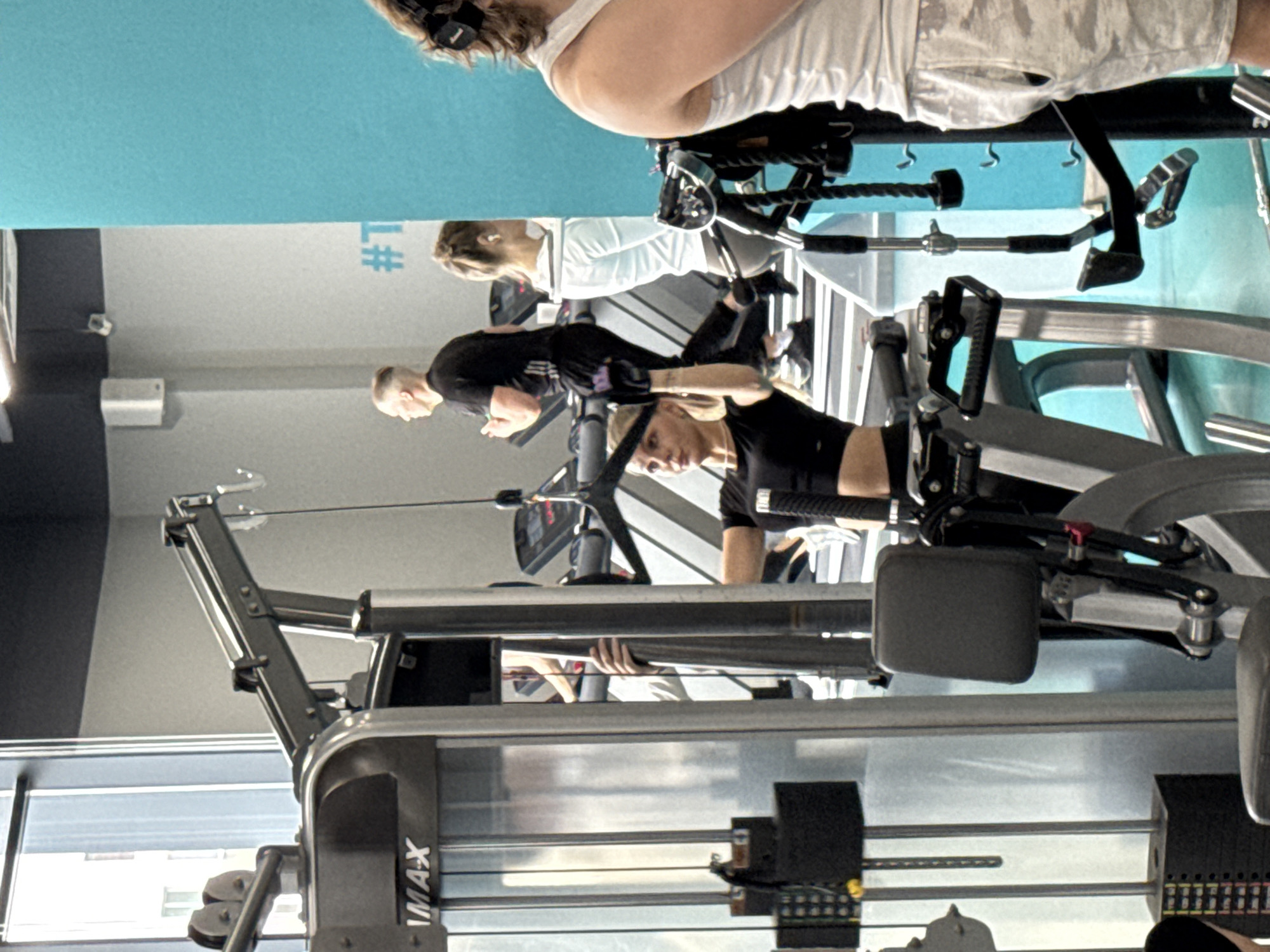 The image shows people exercising in a gym in Yekaterinburg, Russia. A woman with blonde hair wearing a black top is using a cable machine. Another woman in white is on a treadmill, and a man in a black shirt is running nearby. In the foreground, there is a stationary bike being used by someone wearing shorts and headphones. The gym has gray walls and a blue ceiling. There are windows along the left side of the frame.