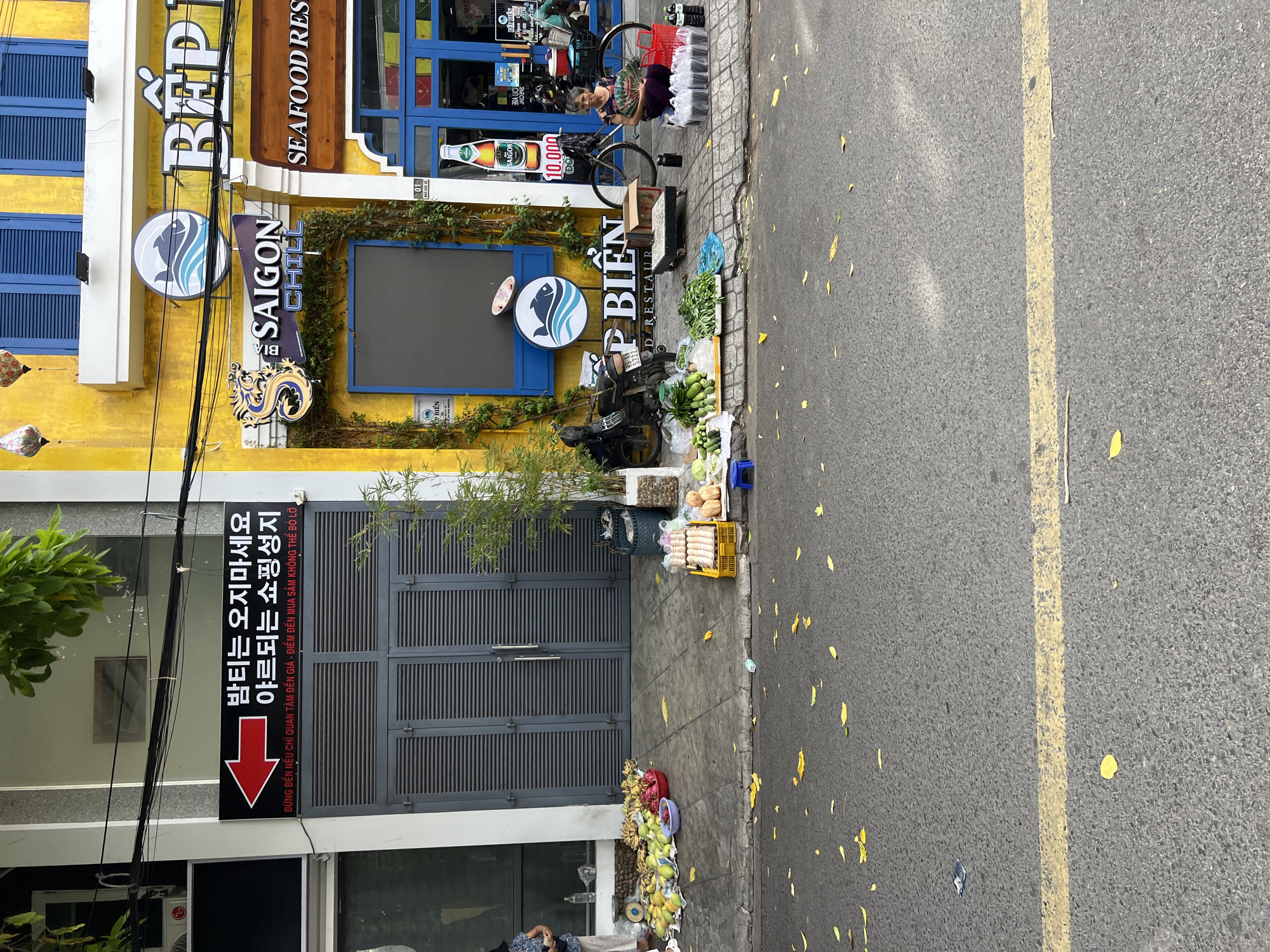 The image captures a street scene in Nha Trang, Vietnam, likely in an urban commercial area. In the foreground, the asphalt road is visible with scattered yellow leaves. A yellow dashed line runs across the road. The sidewalk area is occupied by vendors and shops.
On the left side of the sidewalk, a person is partially visible near a stall displaying various fruits, including bananas and dragon fruit. Next to the fruits, there is a scale and a woman standing. Further back, a large gray door with horizontal slats is visible. A black sign with red text and an arrow pointing left is mounted above the door, with Korean text and Vietnamese text below.
To the right of the door, a brightly painted yellow building dominates the scene. Several signs are displayed on the building. One sign reads "BẾP CHILL SAIGON" with a fish logo. Below this, a sign for "SEAFOOD RESTAURANT" is present. Further to the right, a blue-doored shop is visible with a sign that appears to advertise a beer.
In front of the yellow building, a street vendor is seated on a blue stool, fanning herself with a traditional fan. She is surrounded by bags of goods, possibly food items, and a bicycle is parked nearby. A variety of fresh produce, including cucumbers and leafy greens, are displayed on the sidewalk. A yellow crate contains what appear to be pastries or small breads. Several bottles of liquid are also visible near the vendor. The overall impression is a busy street with both commercial establishments and street vendors operating in close proximity.