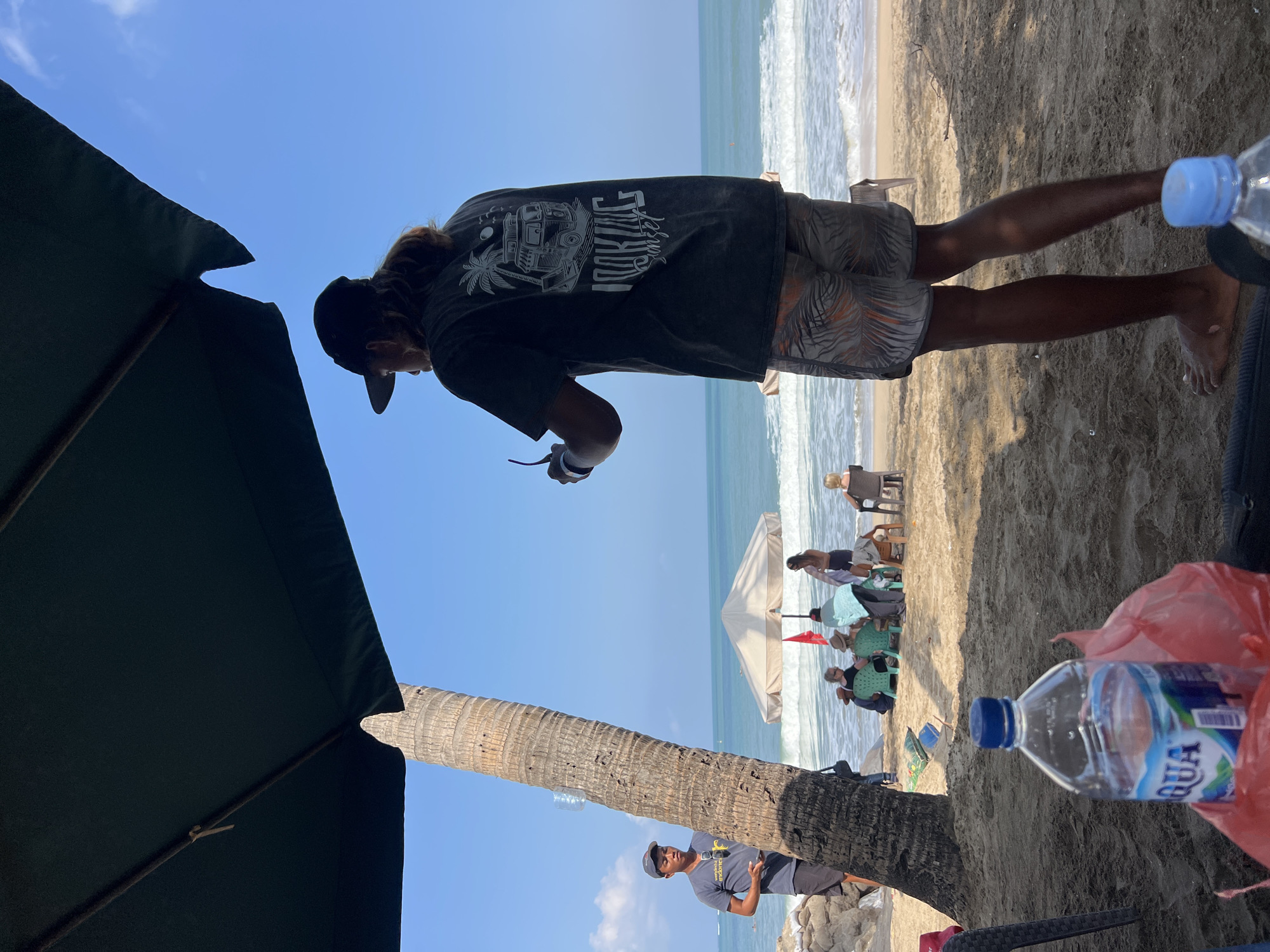 This image depicts a sunny day at Kuta Beach, Bali, Indonesia, with a clear blue sky and gentle waves rolling onto the shore. In the foreground, a person wearing a black t-shirt and patterned shorts stands facing away from the camera, holding sunglasses. A large green umbrella provides shade. To the left, a palm tree stands tall, and another person in a gray t-shirt and shorts is visible. Further back, several people are seated on chairs under a large, light-colored umbrella, enjoying the beach. The sand is a light brown, and scattered beach chairs and a red flag are visible. The ocean stretches out to the horizon, with white surf breaking on the shore.