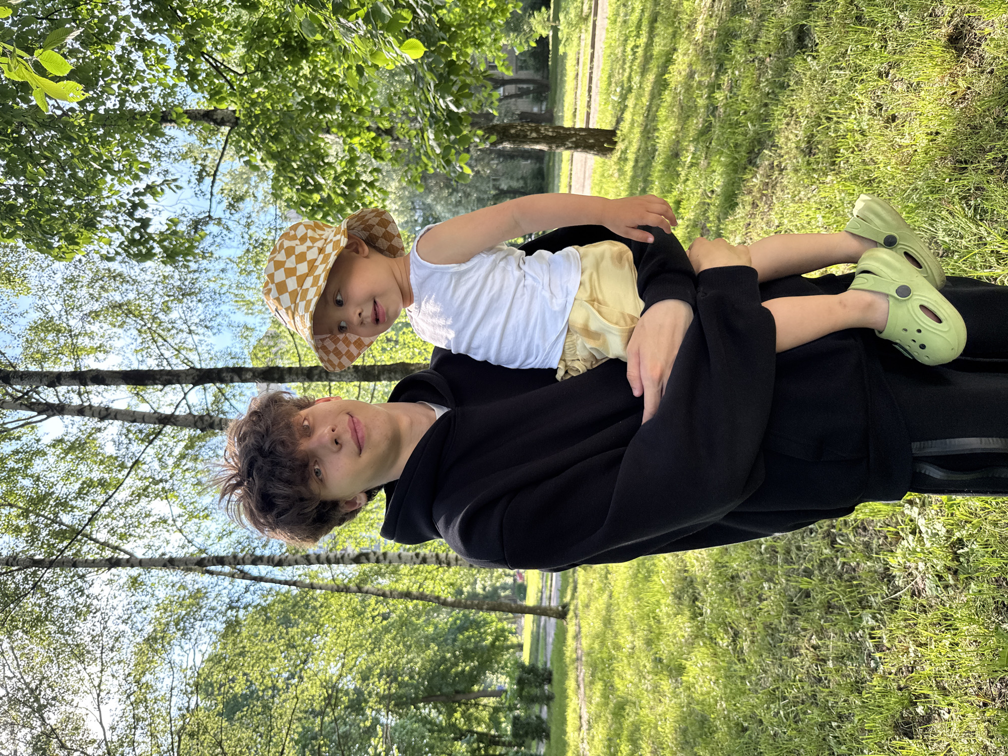 A young man with brown, curly hair, wearing a black hoodie and black pants, stands outdoors holding a toddler. The toddler, wearing a white tank top, yellow shorts, and a checkered bucket hat, is being held in the man's arms, facing to the right. The toddler is wearing light green Crocs. The background consists of a grassy park with numerous birch trees and dappled sunlight. The overall scene suggests a casual outing in a park.