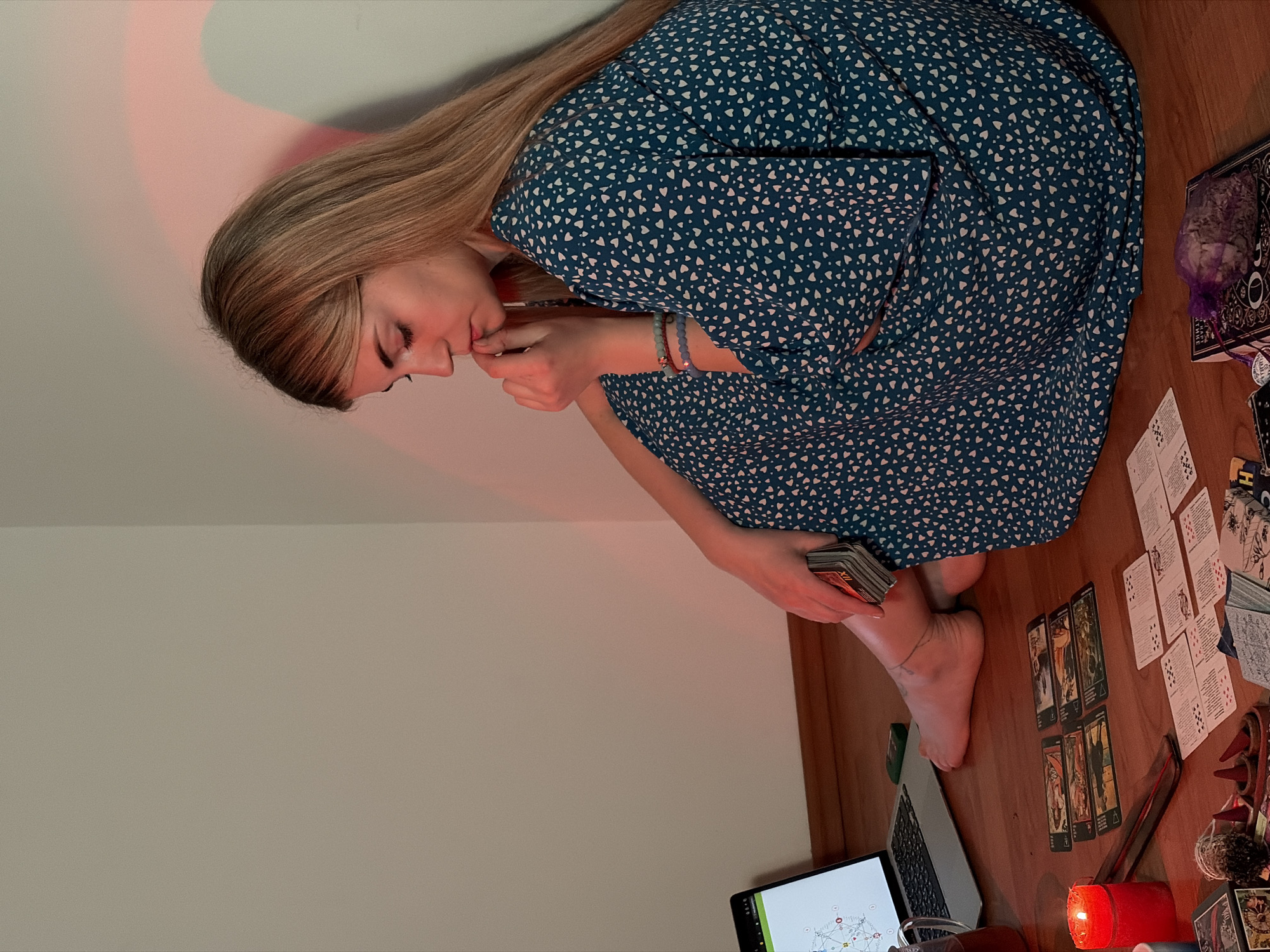 A woman sits on a wooden floor in a room with beige walls, seemingly engaged in a tarot reading. She is wearing a blue dress with white heart patterns and has long, blonde hair. She is holding a deck of tarot cards in one hand, and her other hand is near her mouth. Spread out on the floor around her are various tarot cards, a lit red candle, a laptop displaying a geometric diagram, books, and other objects possibly related to divination. A red glow emanates from the corner near the ceiling. The overall setting suggests a quiet, private space where the woman is focused on performing or consulting tarot readings. There is no information in the image that specifically identifies the location as Budva, Montenegro.