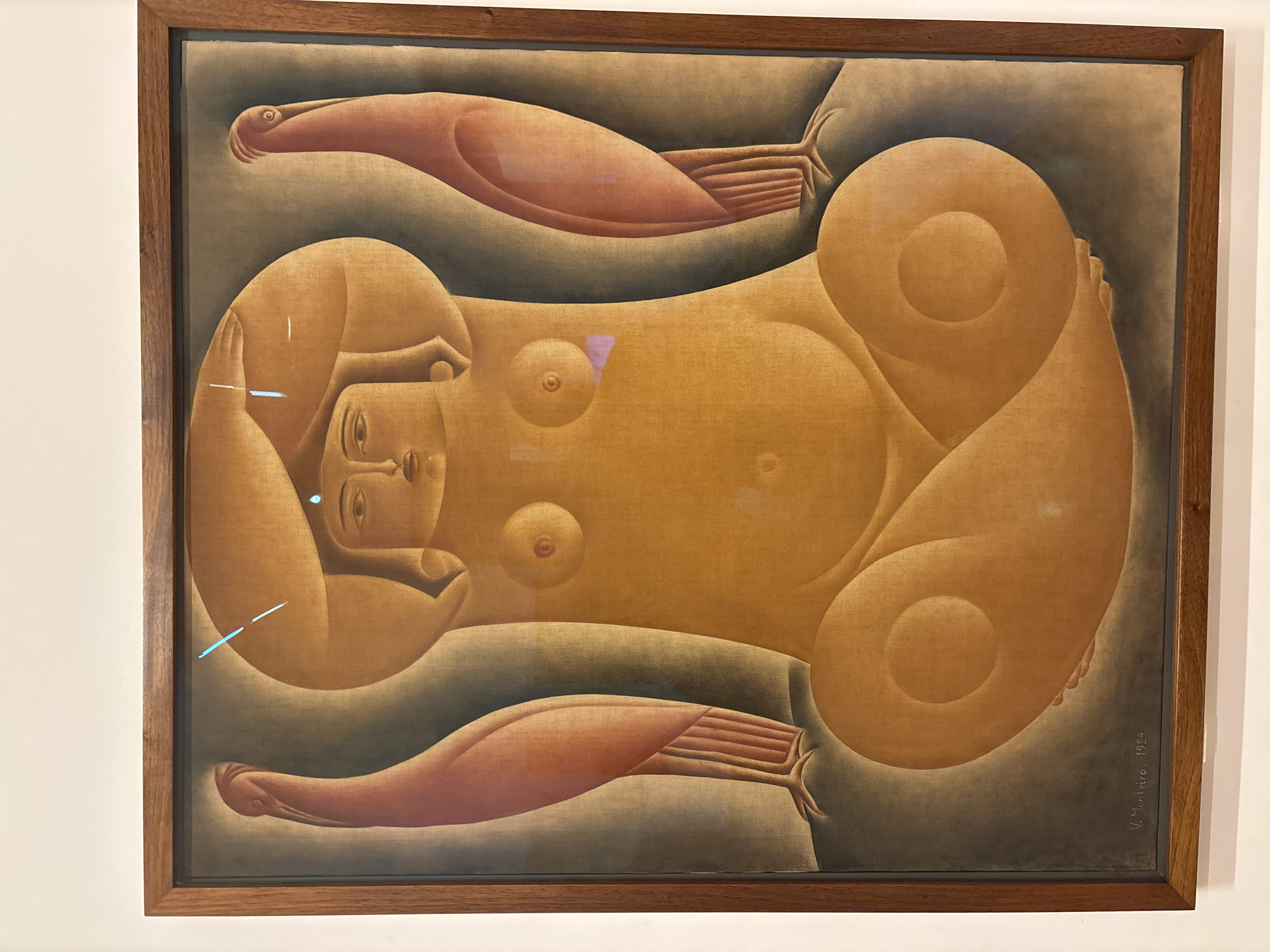 Framed painting titled "Reclining Figure" by Vicente do Rego Monteiro, dated 1924. The painting depicts a stylized, nude female figure lying on her back with arms and legs extending out from her body. The figure is depicted in shades of tan and brown, with simplified, rounded forms. The background is a muted gray. The artist's signature is visible in the lower right corner of the painting. The painting is on display in the Pinacoteca do Estado de São Paulo, Brazil.
