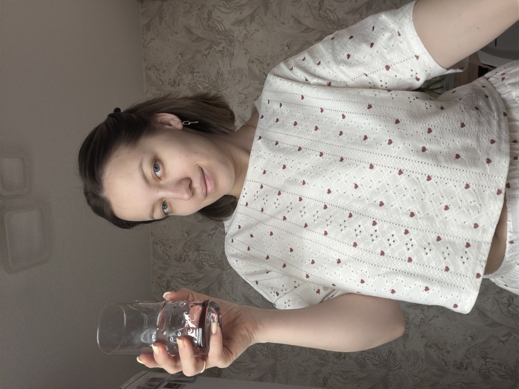 The image shows a woman holding an empty glass. She is wearing a white top with red heart patterns. The background includes a wall with a textured pattern. The location is suggested as Reshetnikovo, Russia. There is no specific information in the image to definitively confirm this location.