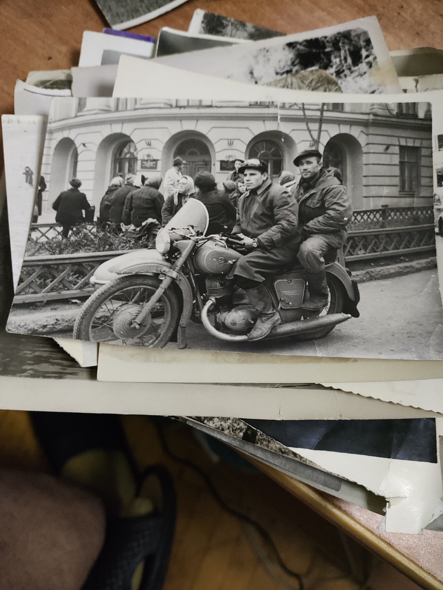 A black and white photograph shows two men riding on a motorcycle. The man in front is wearing a flat cap and a jacket, and the man behind him is also wearing a cap and a jacket. They are both looking forward. The motorcycle is a vintage model with a large front wheel and a visible engine. In the background, there is a building with arched windows and a fence. Several other people are visible in the background, some of whom appear to be standing and watching. The scene is set in a city street. The presence of a vintage motorcycle and the architectural style of the building suggest that the photograph was taken in the mid-20th century. While the image itself does not provide definitive clues to pinpoint the exact location in Izhevsk, Russia, it is plausible to infer that this scene represents a street or public space within the city during that historical period.