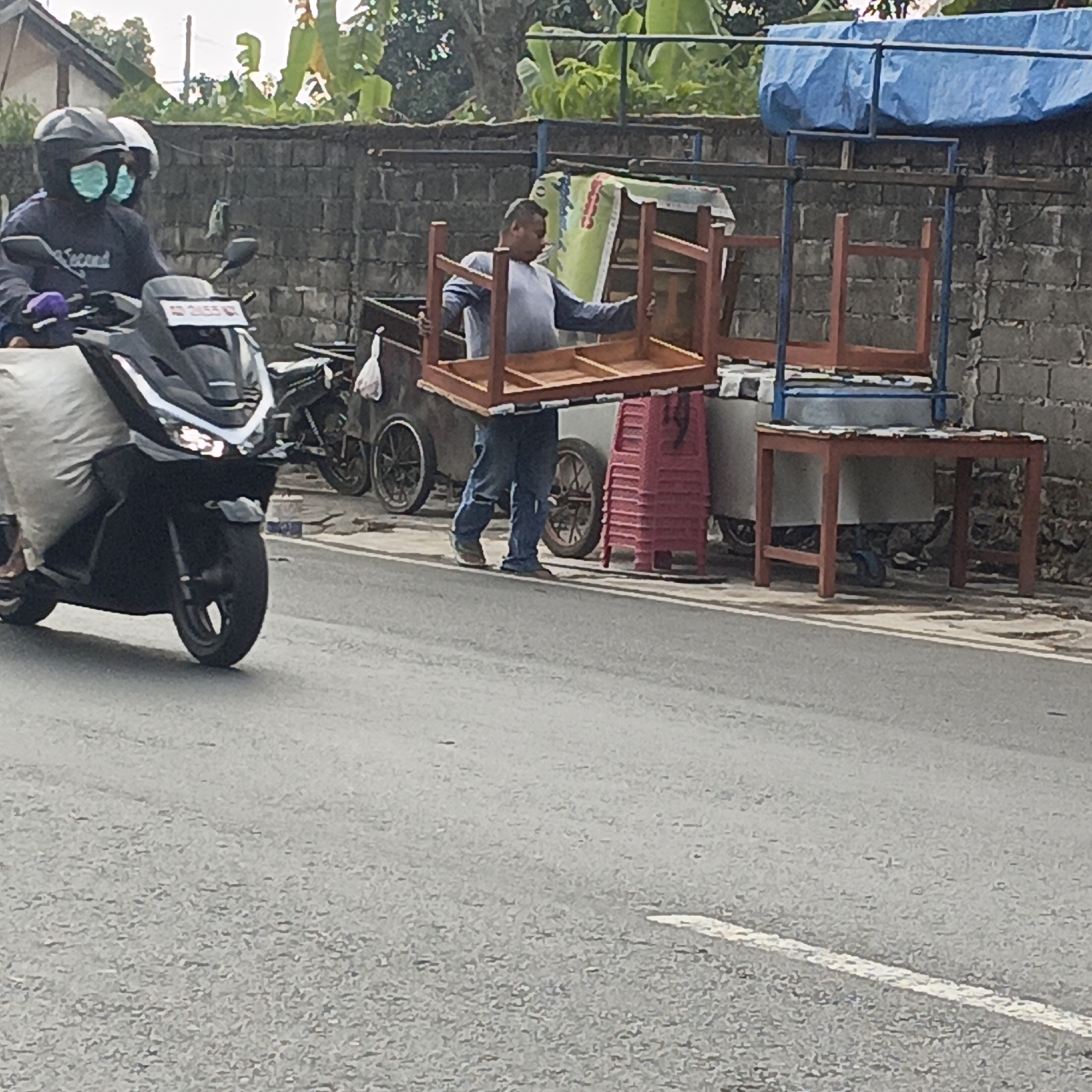 Two individuals wearing masks and helmets are on a motorcycle on a street in Salatiga, Indonesia. One person is driving, while the other is seated behind, carrying a large sack. In the background, a man is lifting a wooden table, possibly for a food stall. There are also other tables, chairs, and carts visible, suggesting a street market or vendor setup. The scene takes place near a brick wall and some greenery.