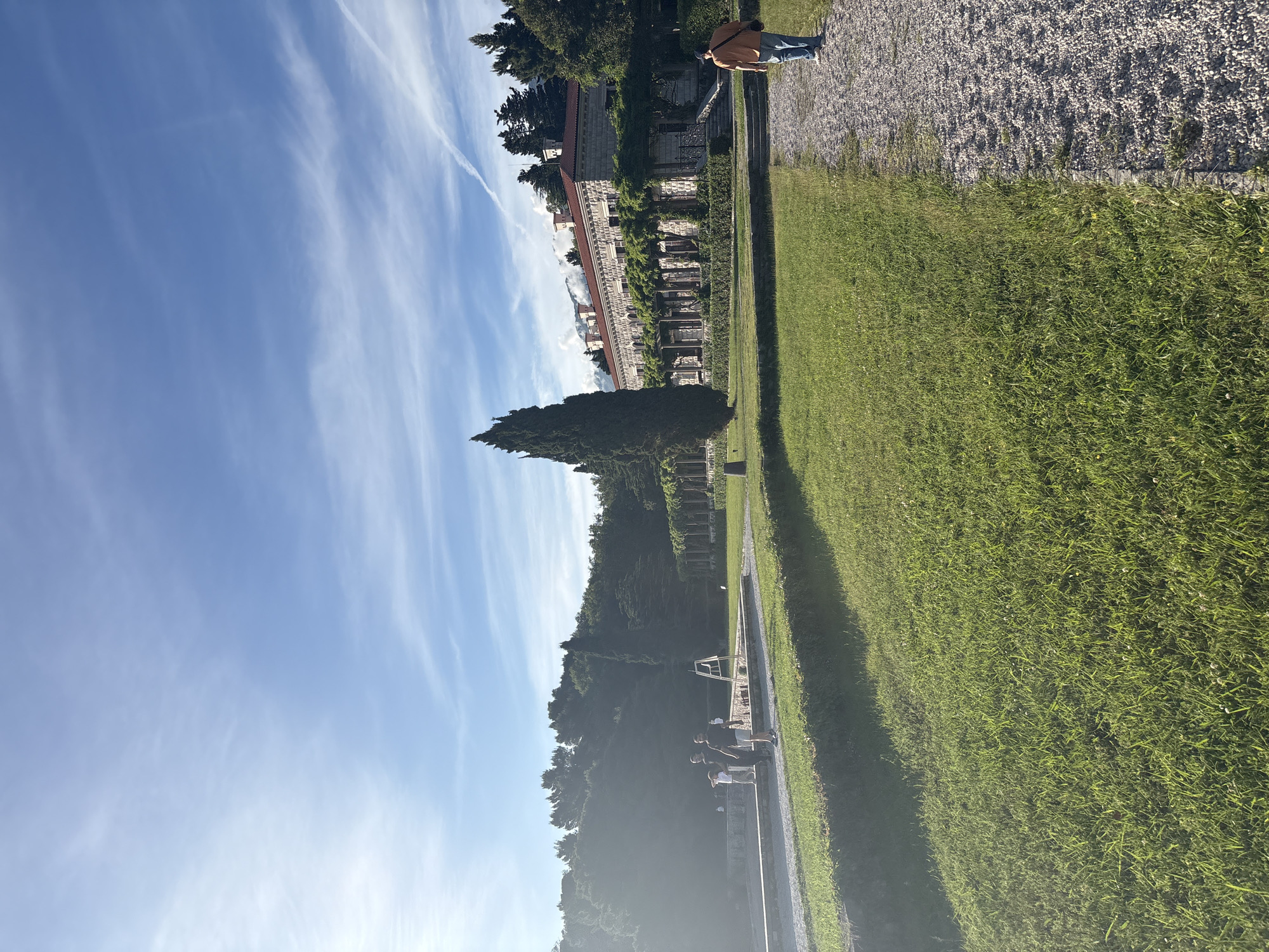 The photograph shows a landscape in Sveti Stefan, Montenegro. The scene is oriented vertically with the sky on the left and the ground on the right. The sky is bright blue with wispy clouds. On the ground, there is a large, well-maintained green lawn. Parallel to the lawn is a walkway lined with trees and vegetation, leading to a structure resembling a building or monument. There is a person standing on a gravel path next to the lawn. In the distance, the outlines of other buildings and trees can be seen against the sky.