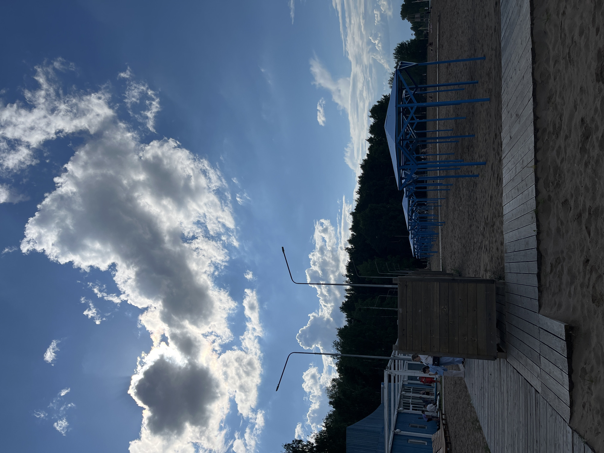 This image depicts a beach area in Chaikovsky, Russia, on a partly cloudy day. In the foreground, a wooden boardwalk leads across a sandy beach. To the left, a light blue building with white trim and an outdoor seating area is visible. Several people are present near this building, some appearing to be relaxing or interacting. A wooden structure, possibly a changing room or shower, stands prominently on the boardwalk. Further into the scene, a row of blue-covered beach shelters with metal supports stretches across the sand. In the background, a dense line of dark green trees forms the horizon, with streetlights visible amongst them. The sky is a vibrant blue, punctuated by large, fluffy white clouds, with some sunlight breaking through.