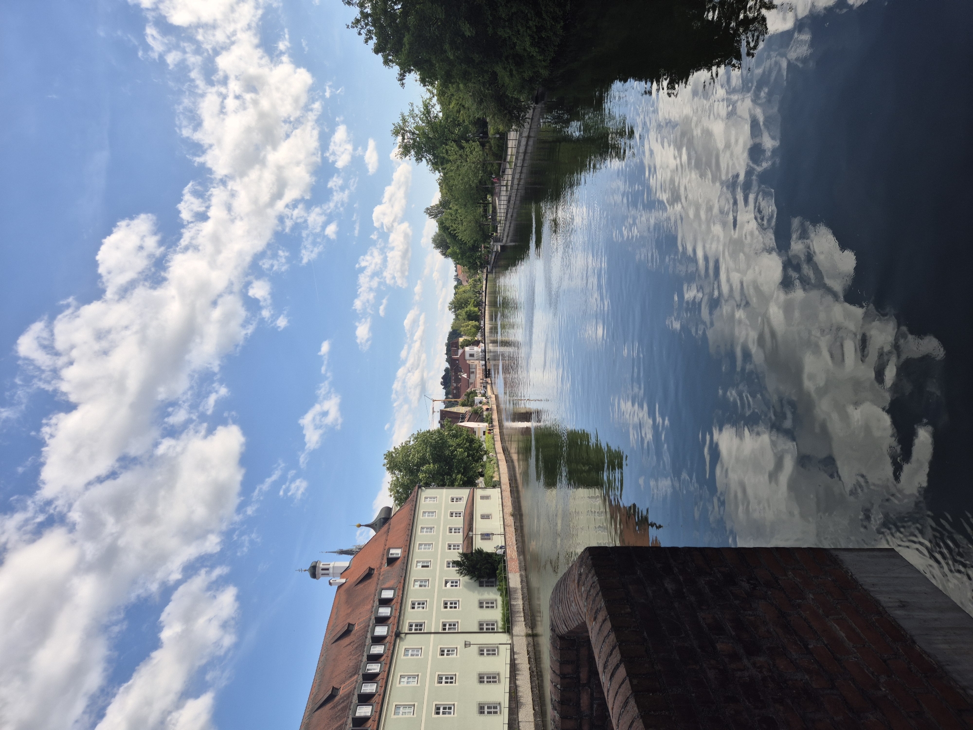 The image captures a scenic view of a canal in Landshut, Germany. The canal, reflecting the sky and surrounding buildings, flows through the city, bordered by trees and pathways. On the left bank, a large, pale green building with a red tiled roof and numerous windows stands prominently. A brick wall in the foreground frames the left side of the canal. On the right bank, a tree-lined promenade with a fence separates the waterway from the urban landscape. In the distance, other buildings and church spires are visible, hinting at the city's architecture. The sky is a vibrant blue, dotted with fluffy white clouds, contributing to the overall picturesque atmosphere. There are no people prominently visible in the scene.