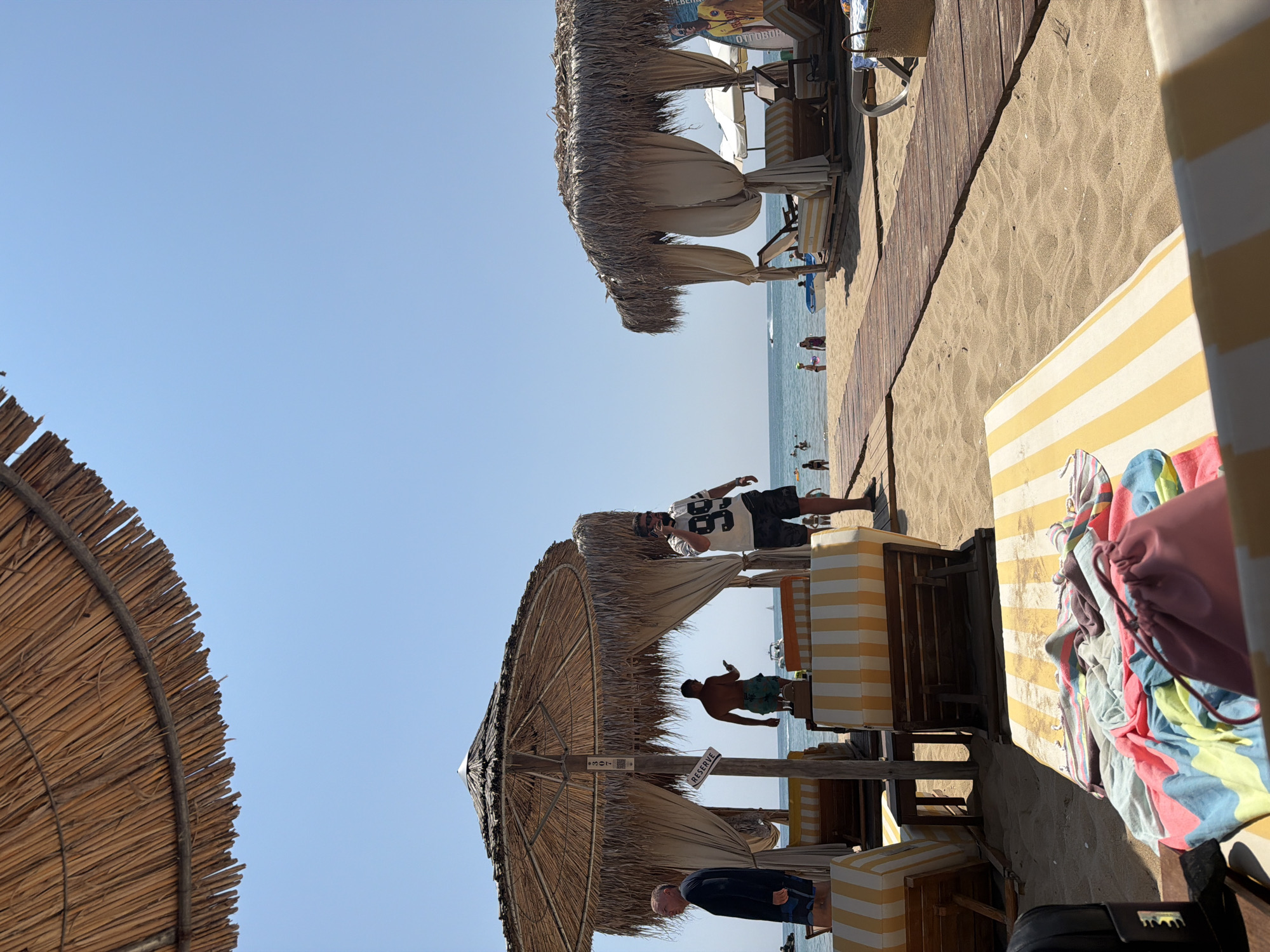 This image depicts a sunny day at a beach resort in Varna, Bulgaria. The foreground features beach chairs with yellow and white striped cushions, some covered with colorful towels and a pink drawstring bag. A wooden walkway extends across the sandy beach towards the sea. In the midground, two thatched-roof cabanas with draped curtains provide shade for lounge chairs. Several people are visible; one man in a white t-shirt with the number "99" is standing near a cabana, holding a phone to his ear. Another man, wearing a dark shirt, stands to the left, and a third man in swim trunks is in the background near the cabana. Further back, the sea is calm, with several people swimming and relaxing. The sky is clear and blue. The overall scene conveys a relaxed beach vacation atmosphere.